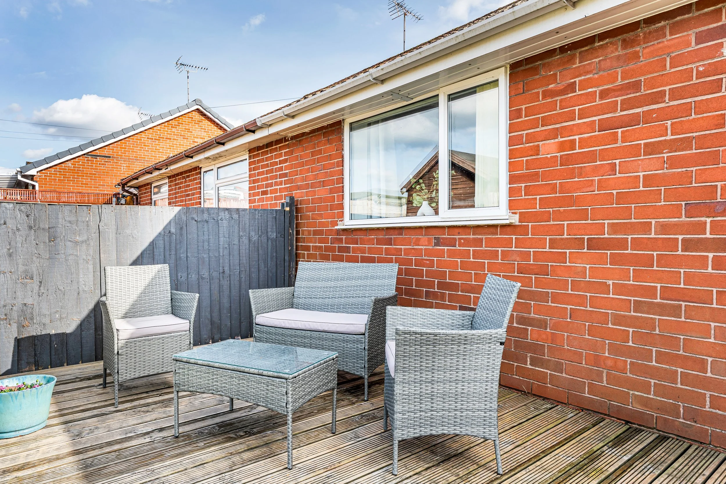 Outdoor patio with wicker furniture including two armchairs, a loveseat, and a glass-top table, on a wooden deck next to a brick house with a window. A black fence and a potted plant are also visible.