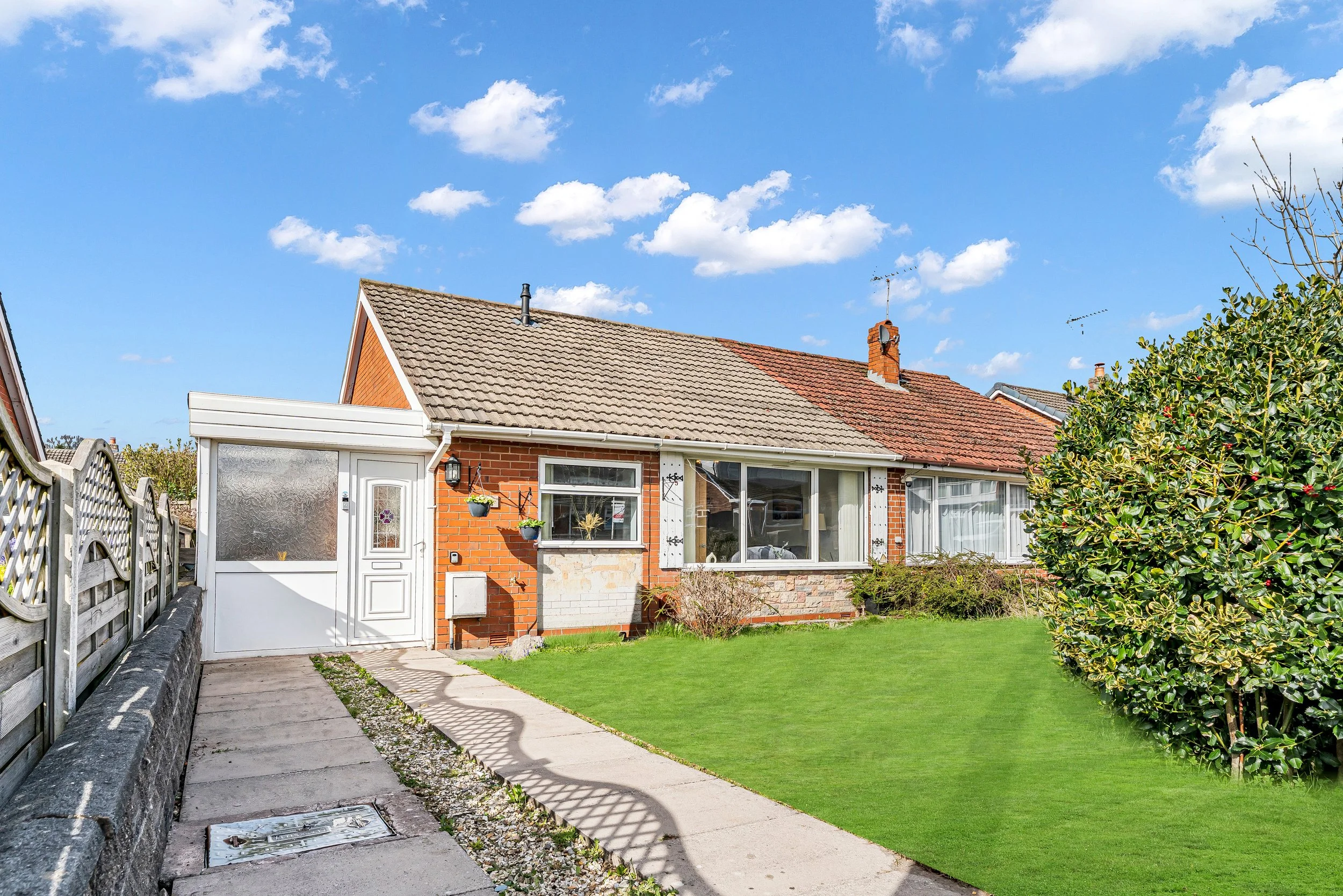 Front view of a single-story brick house with a sloped roof, surrounded by a green lawn and bushes. There is a paved walkway leading to the front door, which has a white door with frosted window. The house has large windows with white frames and hang