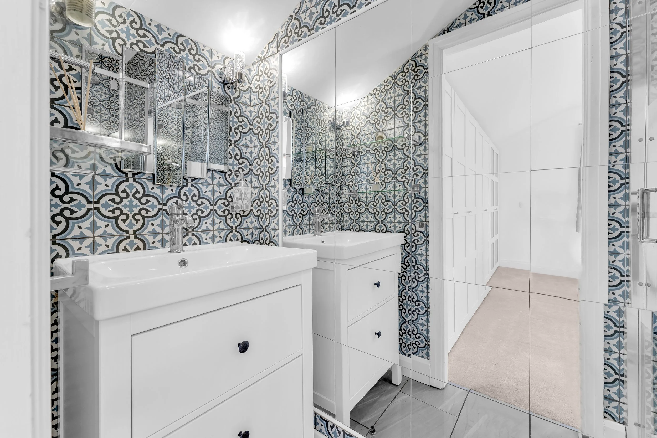 A bathroom with patterned blue, gray, and white tiles on the walls, two white sinks, and mirrored cabinets. There are shelves with glass items and a small container in the mirror. A large mirror reflects the room, and a doorway with a beige carpet is