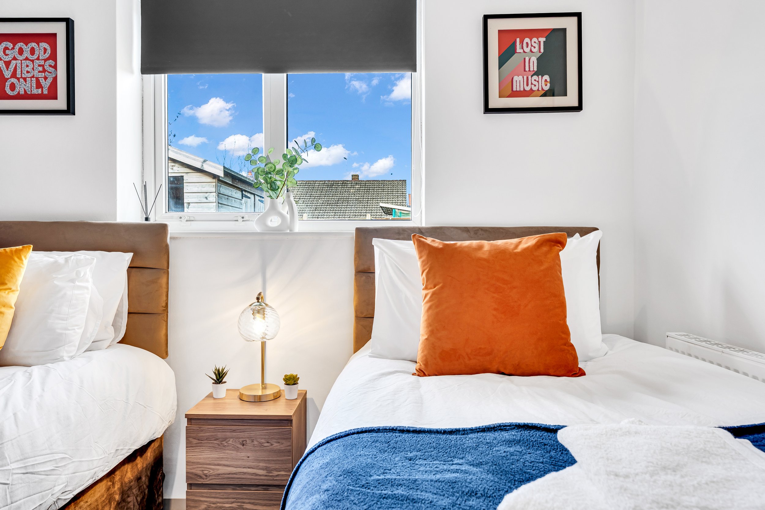 Bedroom with two beds, one with an orange pillow, a wooden nightstand with a lamp and small potted plants, window with a view of blue sky and rooftops, framed wall art, and a white wall.