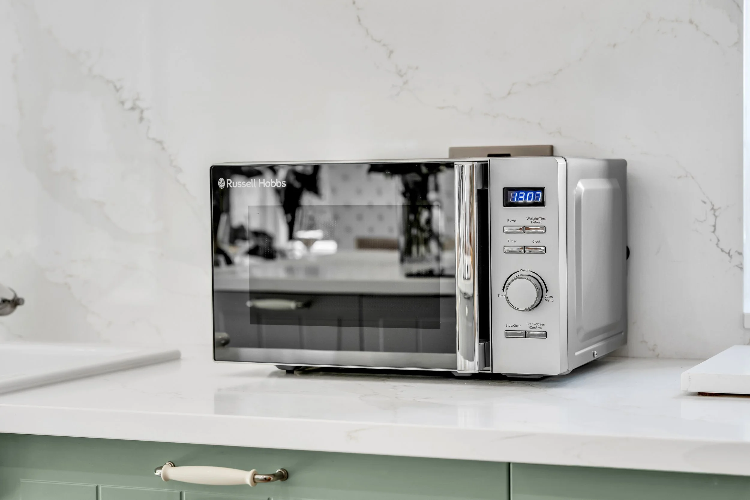 A modern microwave oven with a digital display on a kitchen countertop, reflecting the kitchen interior.