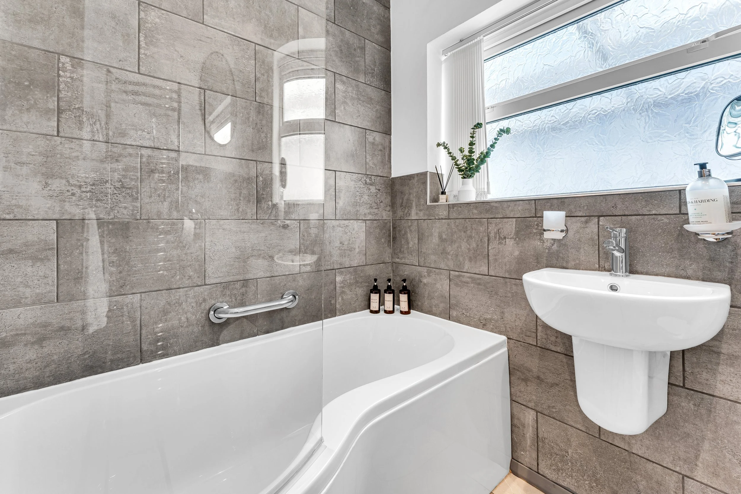 Bathroom with grey tiled walls, a bathtub with a glass shower screen, a small curved white sink, and a frosted window with a potted plant.