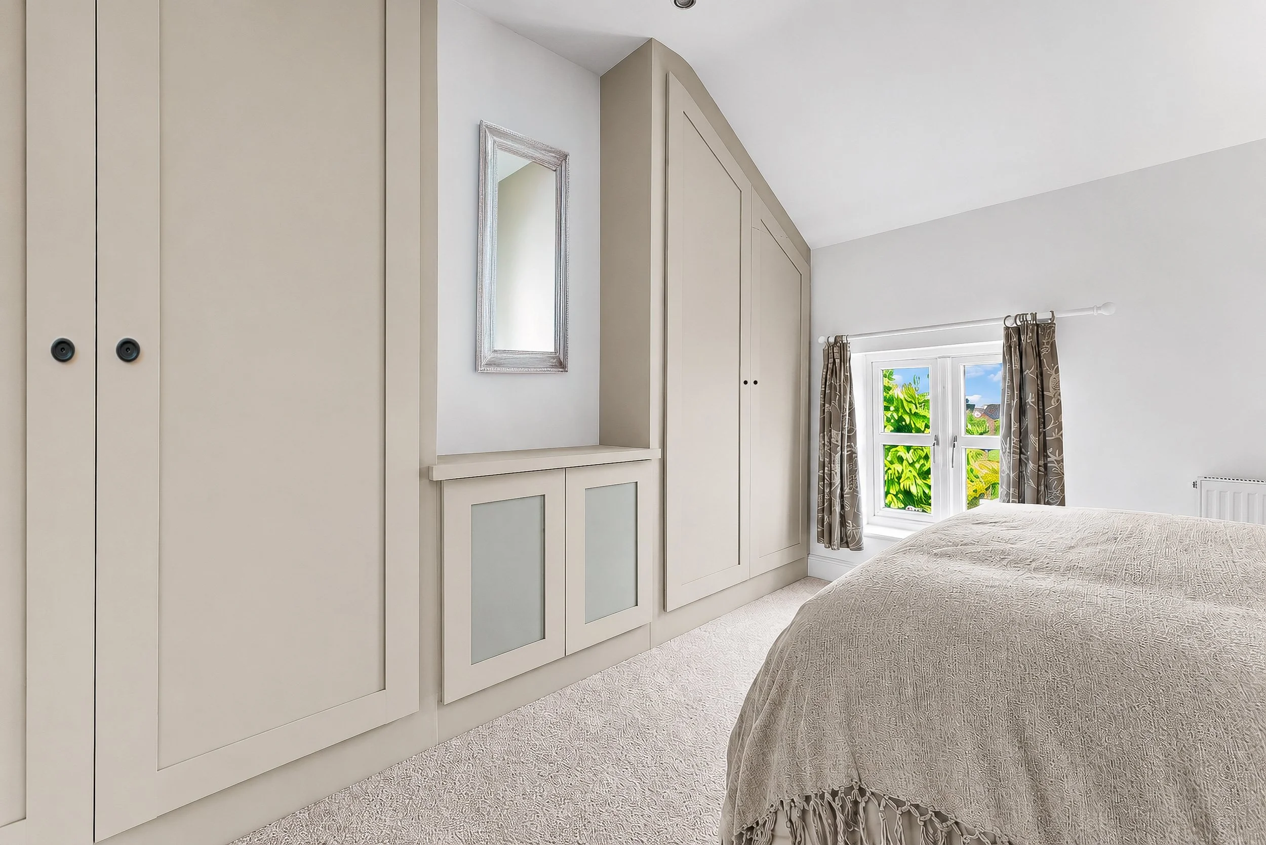 A bedroom with a beige bedspread, built-in white wardrobes, a window with patterned curtains, and a wall mirror. The room has a sloped ceiling and a beige carpeted floor.