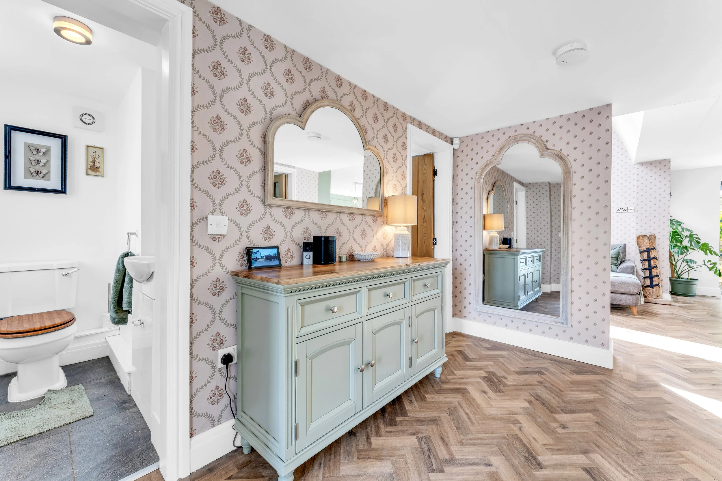 Interior view of a home with patterned wallpaper, a mirror, a sideboard, and a living area with a plant and window.