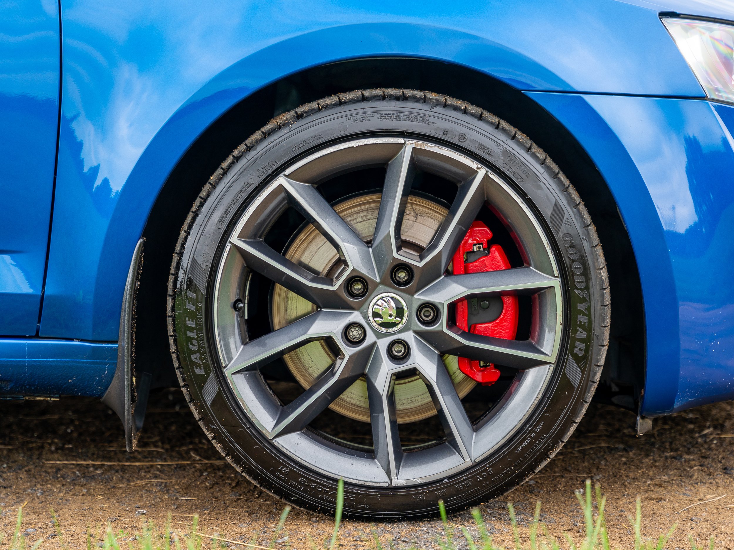 Close-up of a blue car's front wheel with a black and silver rim, red brake caliper, and tire, on a dirt ground with some grass.