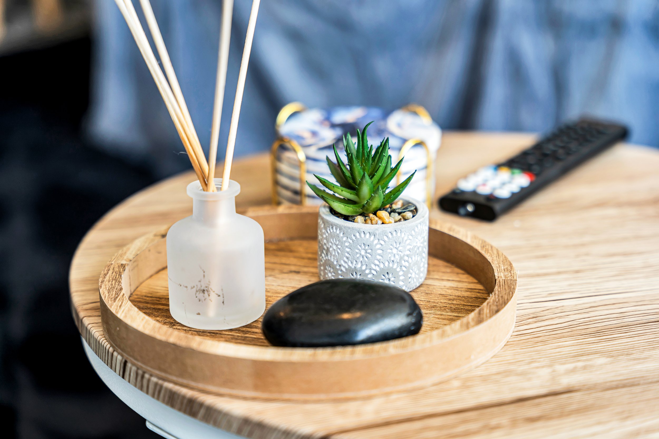 Decorative wooden tray on a round wooden table holding a white frosted glass vase with wooden sticks, a potted succulent plant, a smooth black stone, a wrapped gift box, and a remote control.