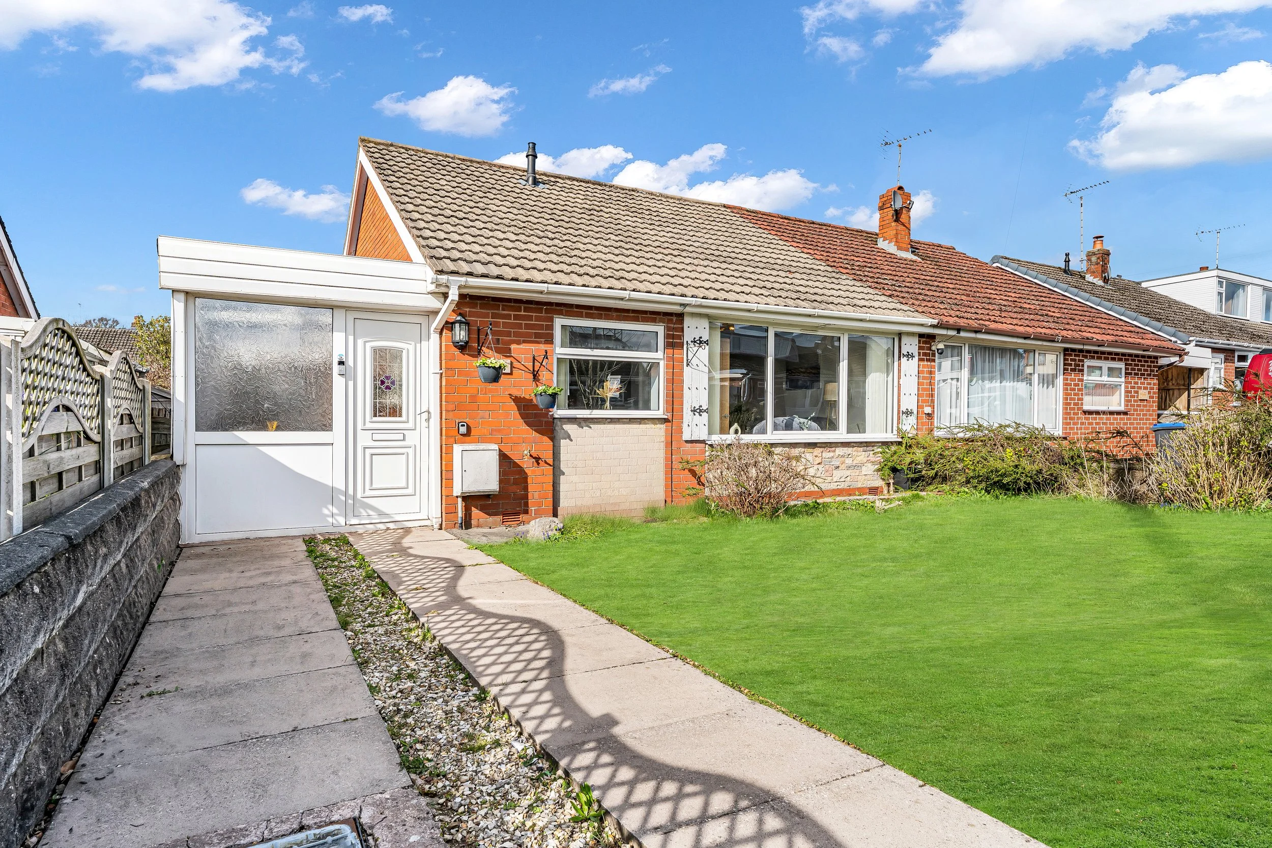 Front view of a single-story brick house with a tiled roof, large front window, and a white door, with a green lawn, concrete walkway, and blue sky with clouds.