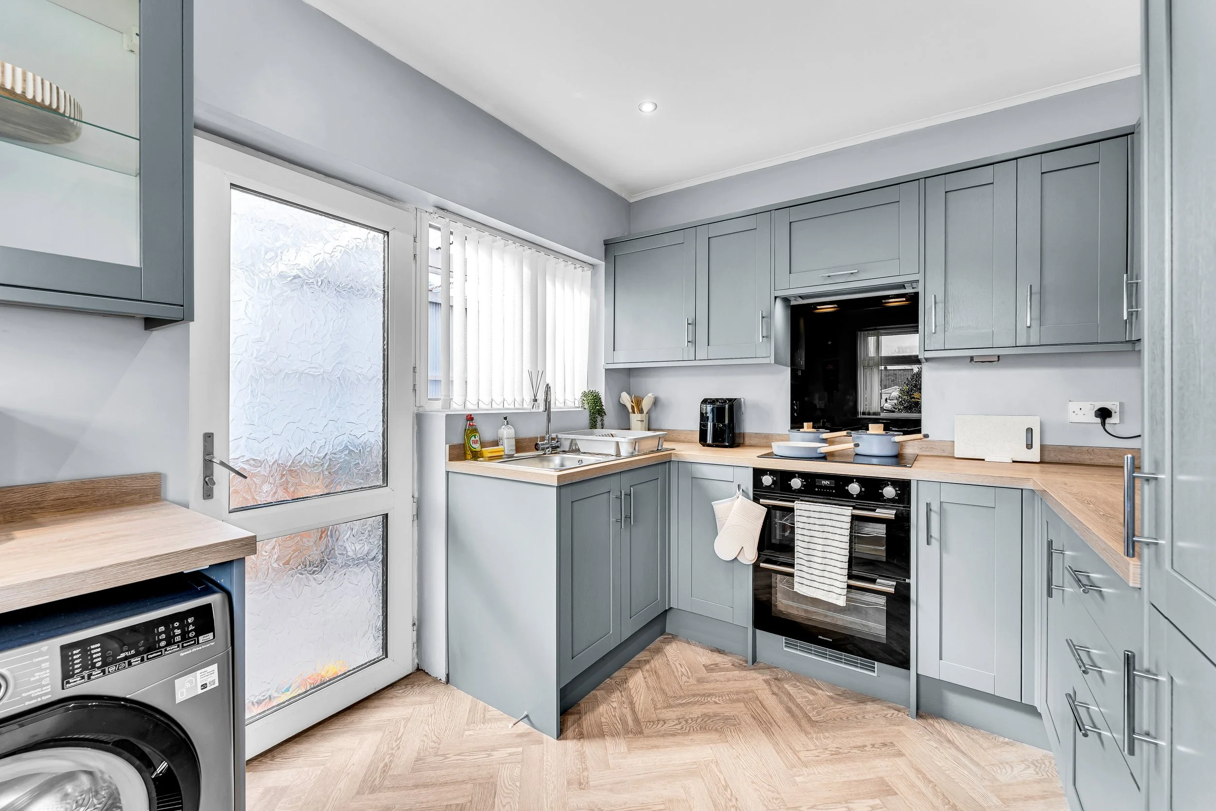 Modern kitchen with light gray cabinets, wooden countertops, a black oven, a washing machine, and a frosted glass door leading outside, illuminated by natural light from a window with vertical blinds.