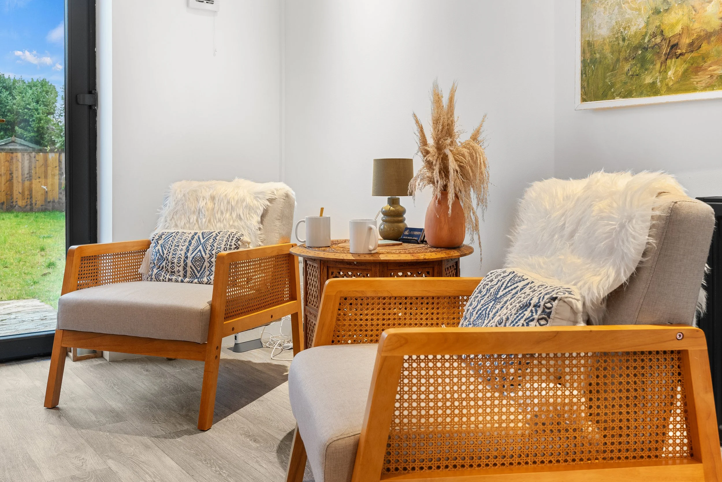 A cozy living room with two beige armchairs with white furry throws and patterned pillows, a round wooden table with a brown vase and pampas grass, a tan table lamp, and two white mugs. A large picture hangs on the wall, and a sliding glass door shows a green backyard outside.