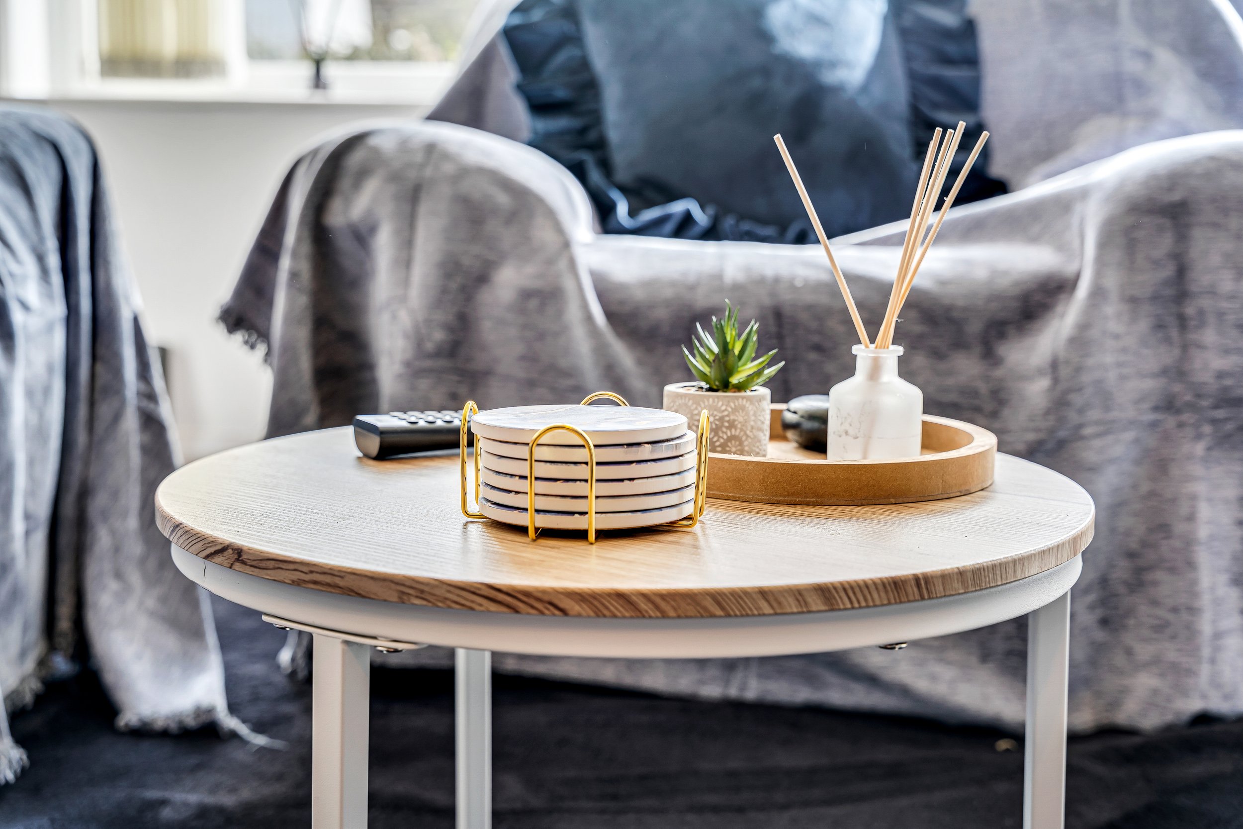 A round wooden coffee table with a white metal frame holding a small potted succulent plant, a set of coasters with gold accents, a remote control, and a white container with reed diffusers. In the background, a gray couch and a blanket are visible.