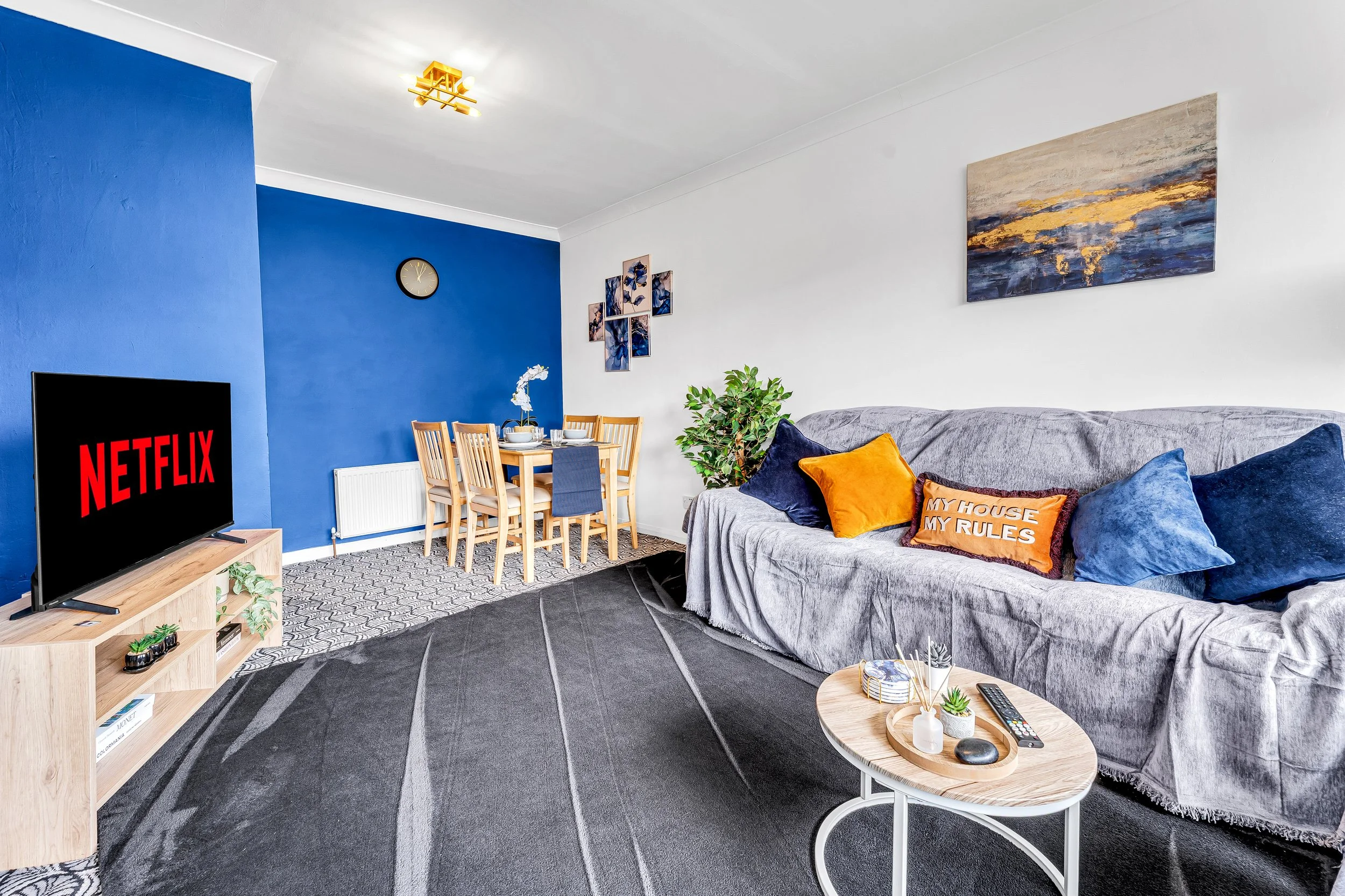 Living room with a gray sofa, blue and orange cushions, a small round coffee table, a TV with a black screen displaying Netflix, a dining table with six wooden chairs, a green plant, art on the walls, and a large window with white trim.