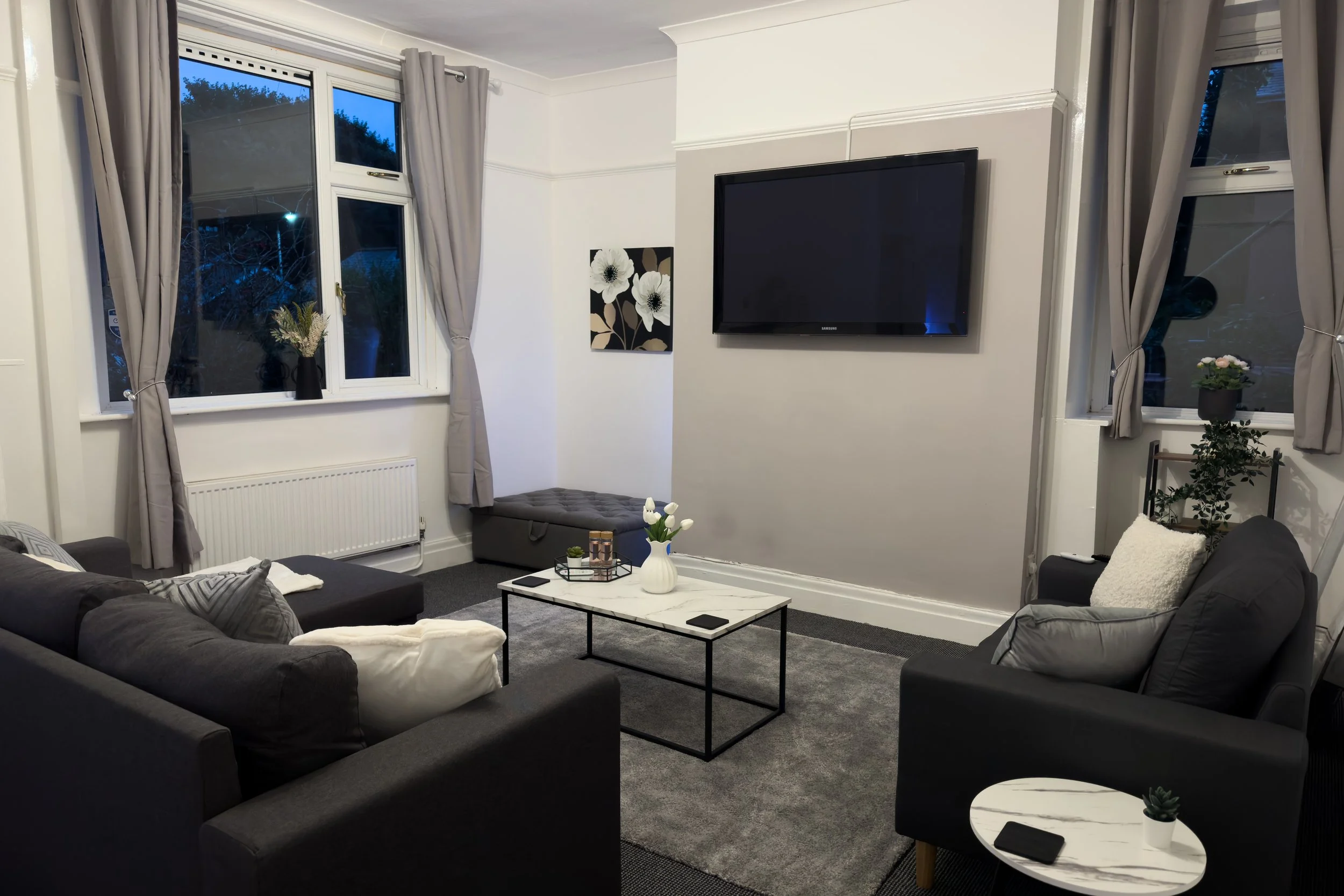 Living room with gray sofa and armchair, white coffee table with a vase of white flowers, patterned pillows, a wall-mounted large TV, and windows with gray curtains, evening outside.