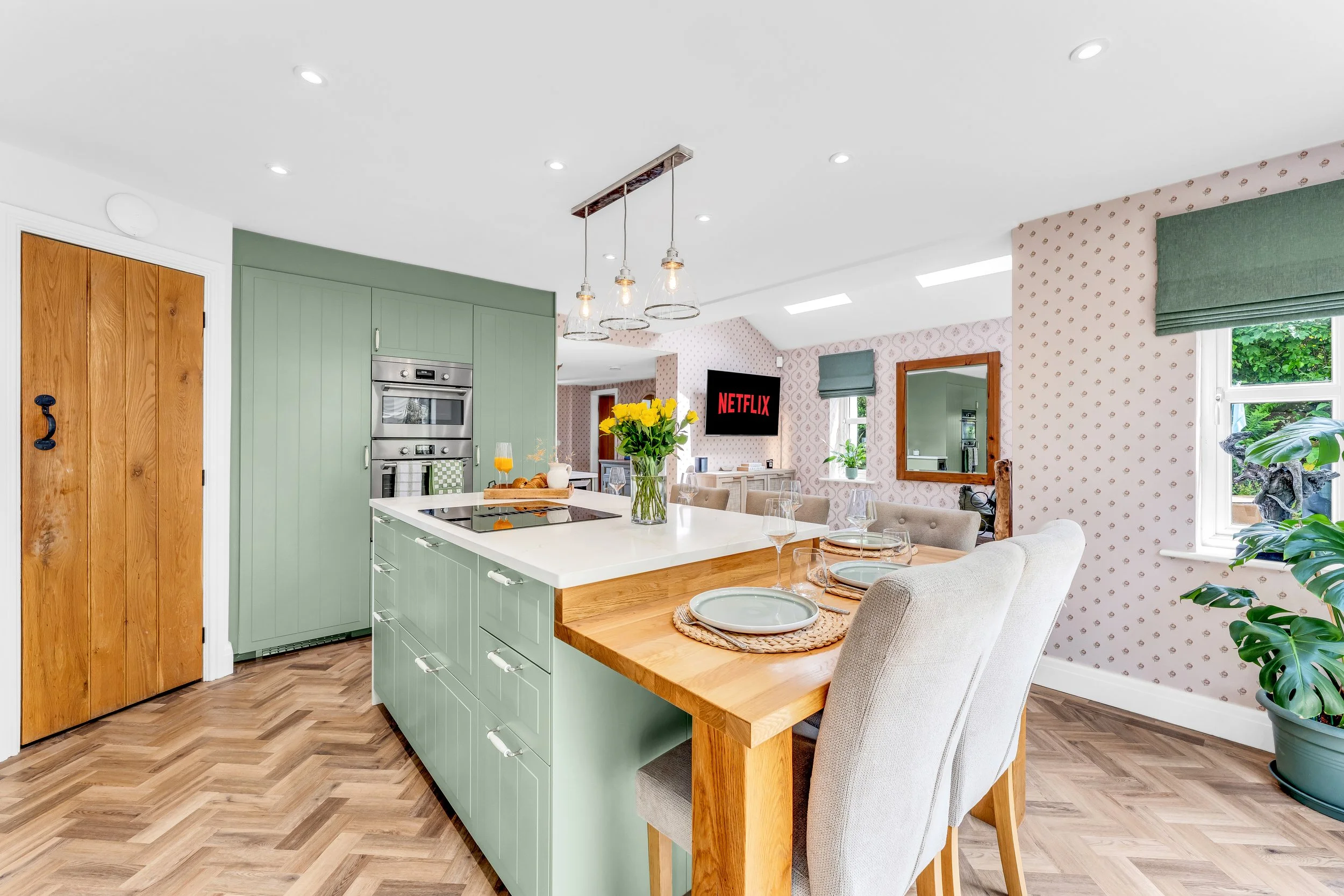 Bright and cozy kitchen and dining area with a green kitchen island, wooden accents, and a dining table set with plates and glasses. A large window offers greenery outside, and a TV showing Netflix is visible in the living space.
