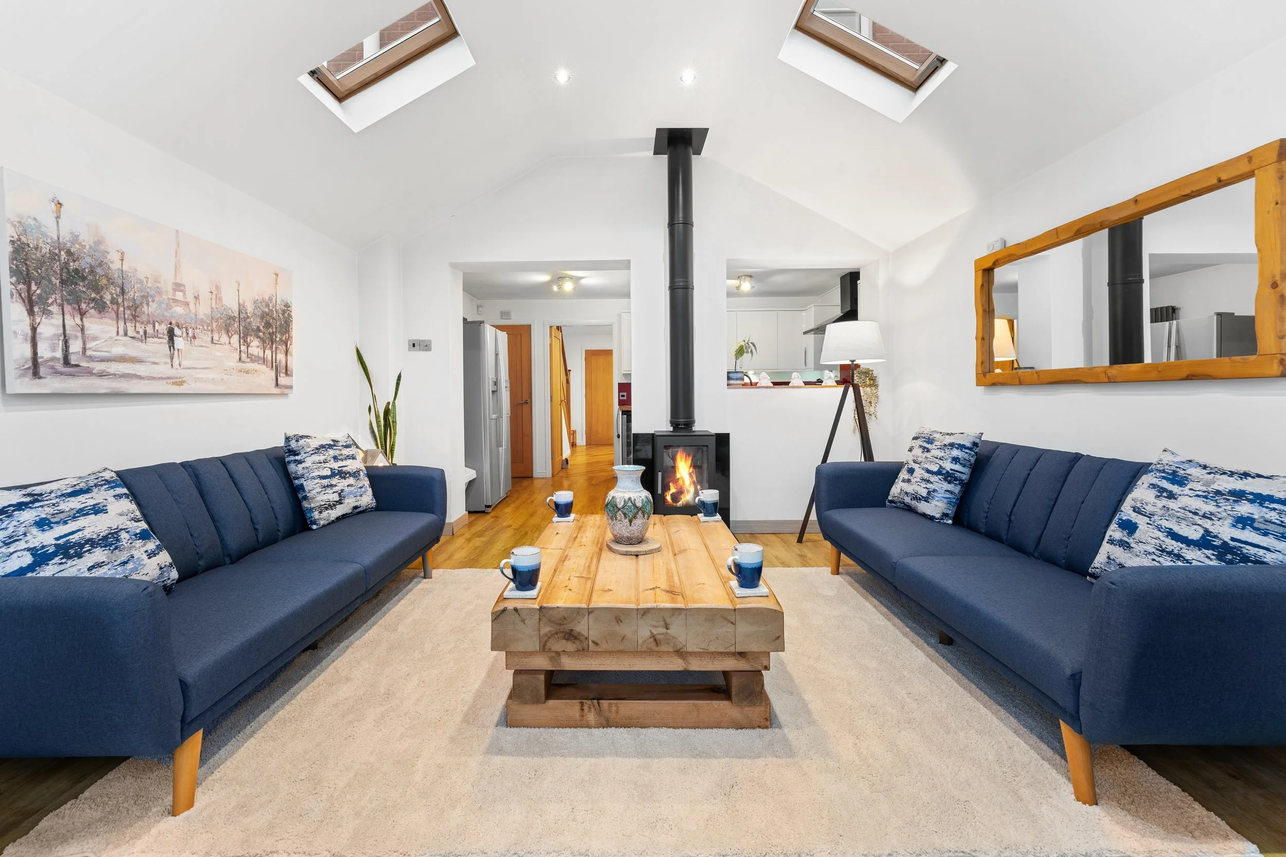 Living room with two blue sofas, a wooden coffee table with six cups, a fireplace, skylights, a large mirror, and a painting of a park scene on the wall.