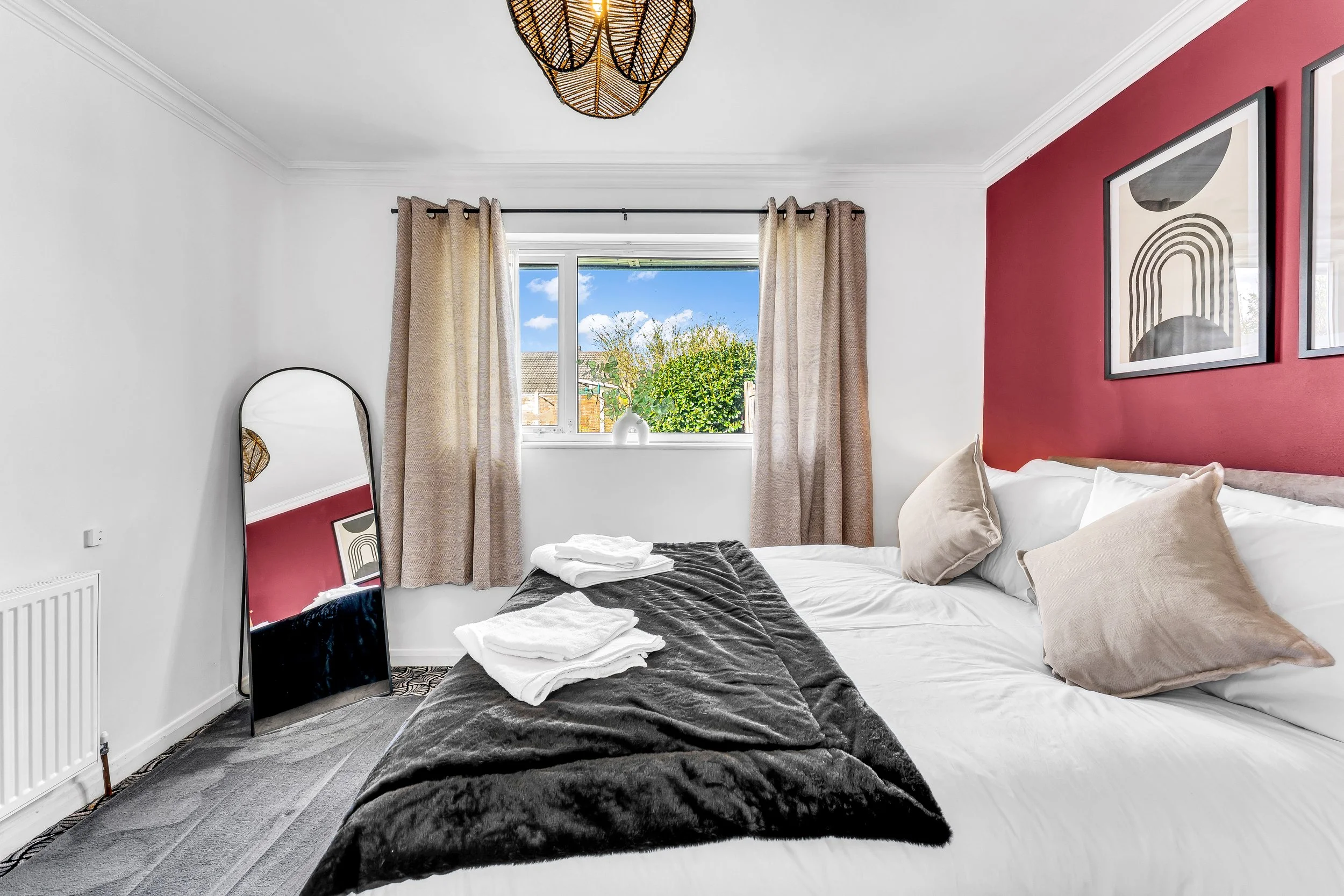 A neatly made bed with white and beige pillows, a black throw blanket, and folded white towels on top, in a bedroom with a window showing a sunny day outside, beige curtains, modern framed art on the wall, and a standing mirror leaning against the wa