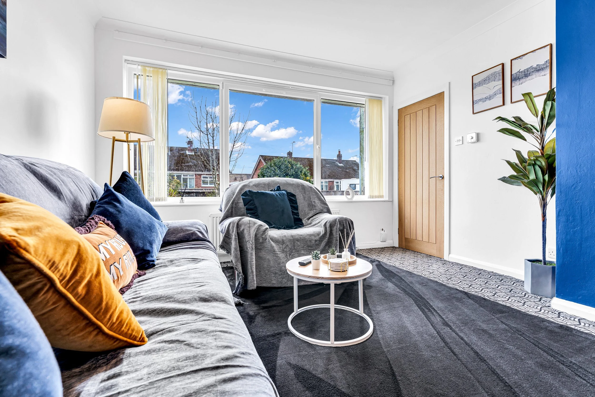 Living room with large window, gray couch with colorful pillows, armchair with dark cushion, round coffee table with decorative items, potted plant, wooden door, white walls, framed artwork, patterned gray and white carpet.