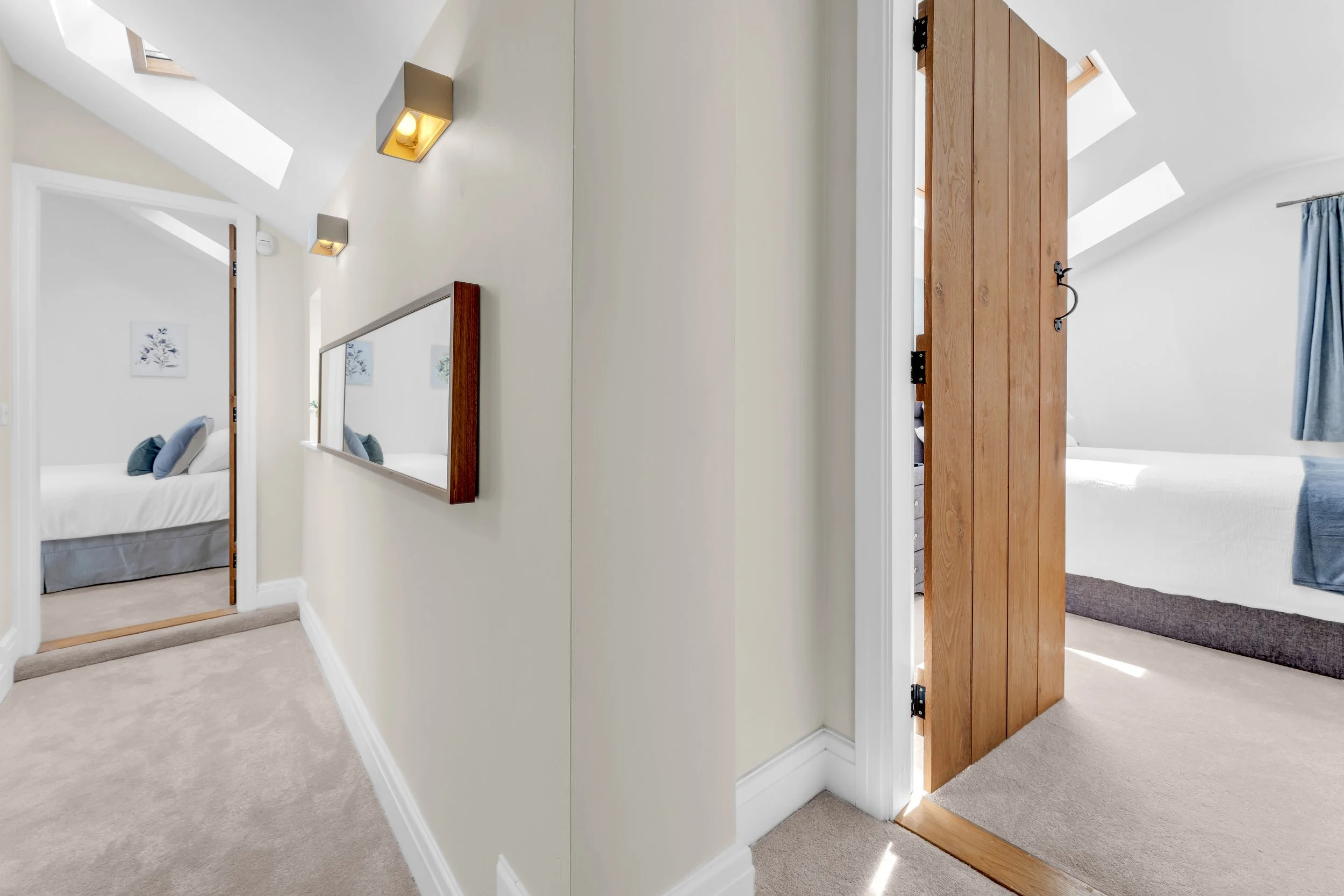 Interior view of a modern house with a bedroom visible through an open wooden door. The hallway has white walls, beige carpet, and a large mirror on the wall. There are two skylights in the sloped ceiling, allowing natural light to fill the space.