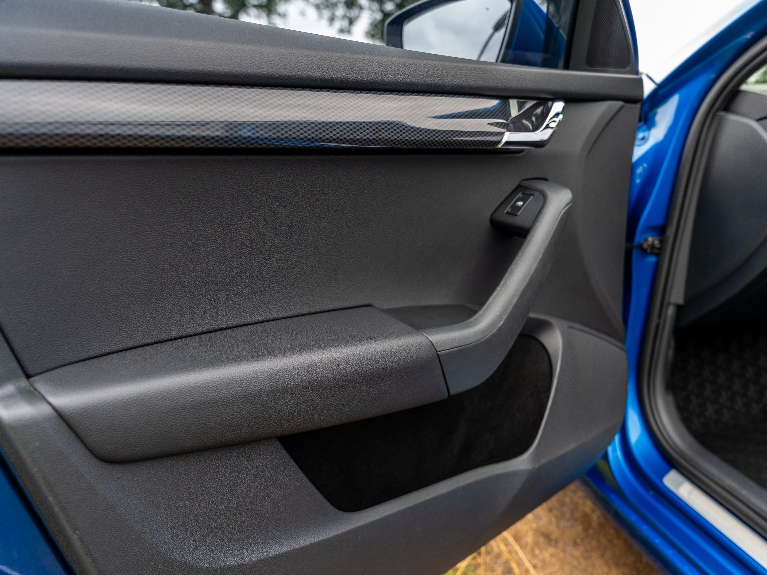 Close-up of a car door interior showing a black door handle with a window control button, blue exterior of the vehicle, and part of the black interior dashboard.