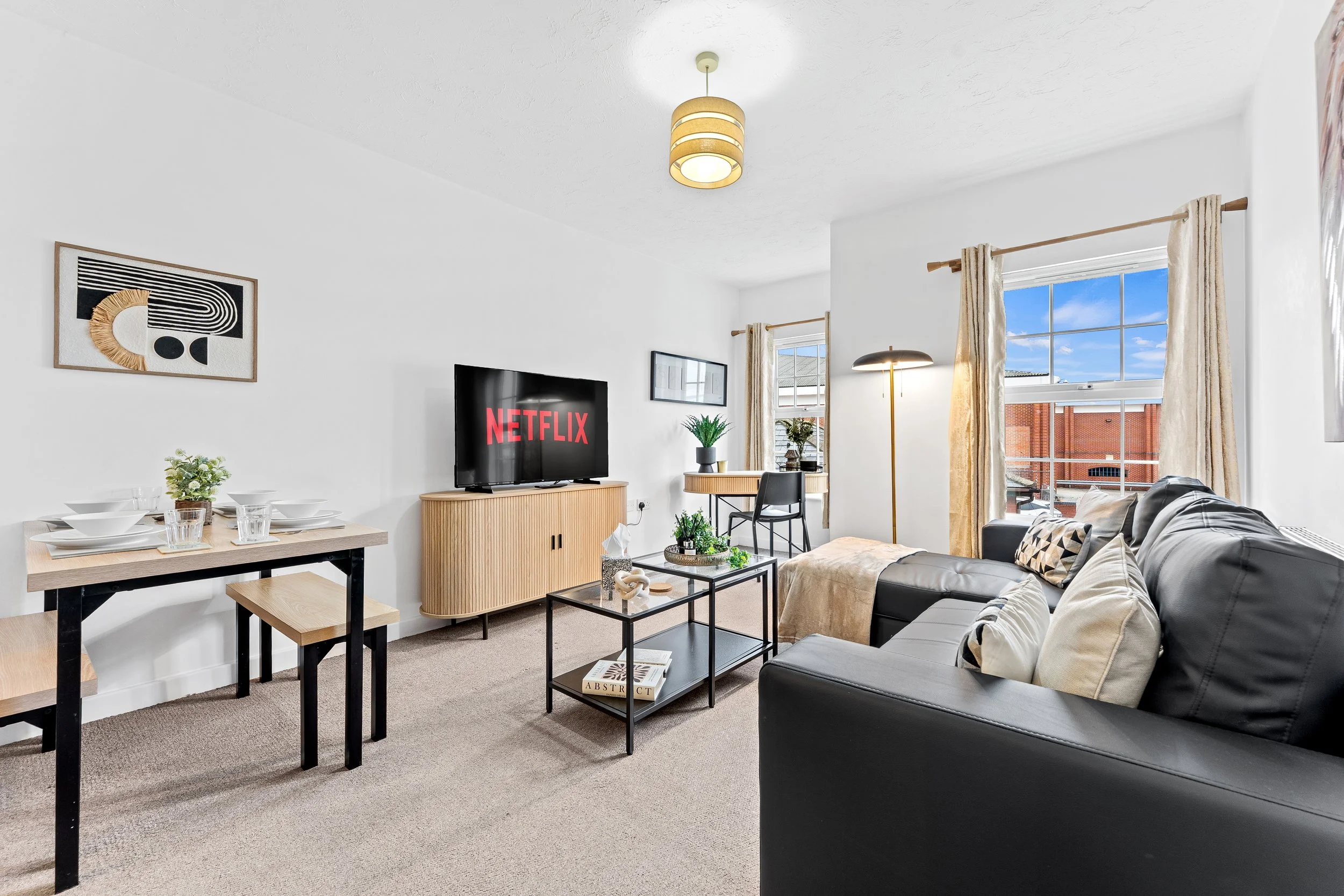 Living room with beige carpet, black leather sofa with decorative pillows, coffee table with books and plants, wall-mounted TV displaying Netflix, side table with potted plants, dining area with table and chairs, large windows with light curtains, and modern lighting fixtures.
