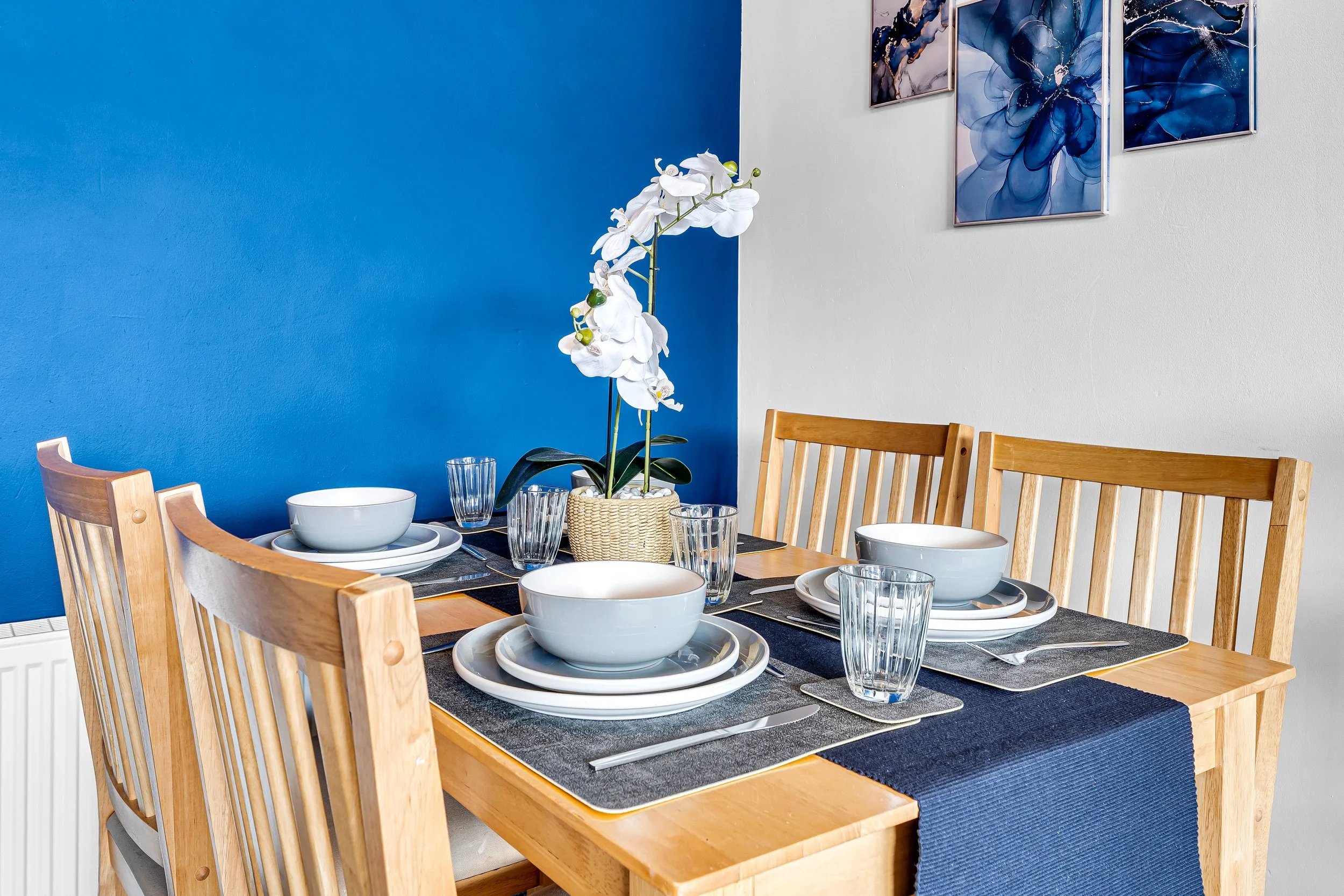 Dining table set with bowls, plates, glasses, and a potted white orchid, in a room with a blue and white wall, and framed blue floral artwork.