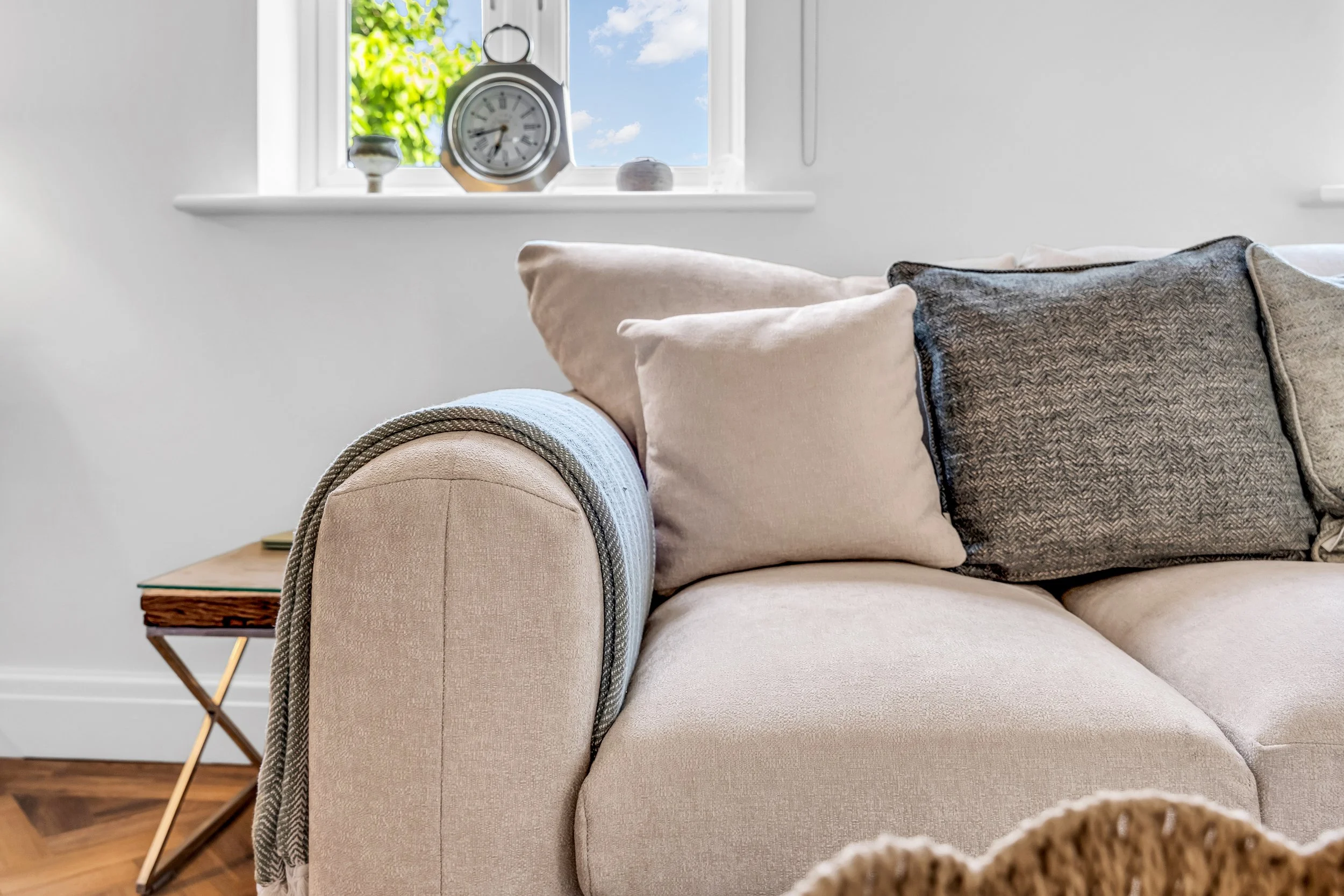 A beige sofa with multiple pillows, including light and dark gray textures, in a room with a window showing blue sky and green trees outside. A wooden side table and a decorative clock are visible on the windowsill.