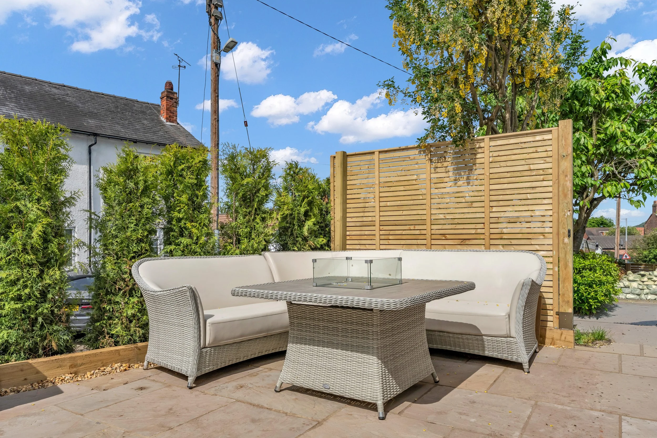 An outdoor patio area with a wicker sofa and matching chairs set around a rectangular table, with a large wooden privacy fence, trees, and a house with a chimney in the background, under a blue sky with some clouds.
