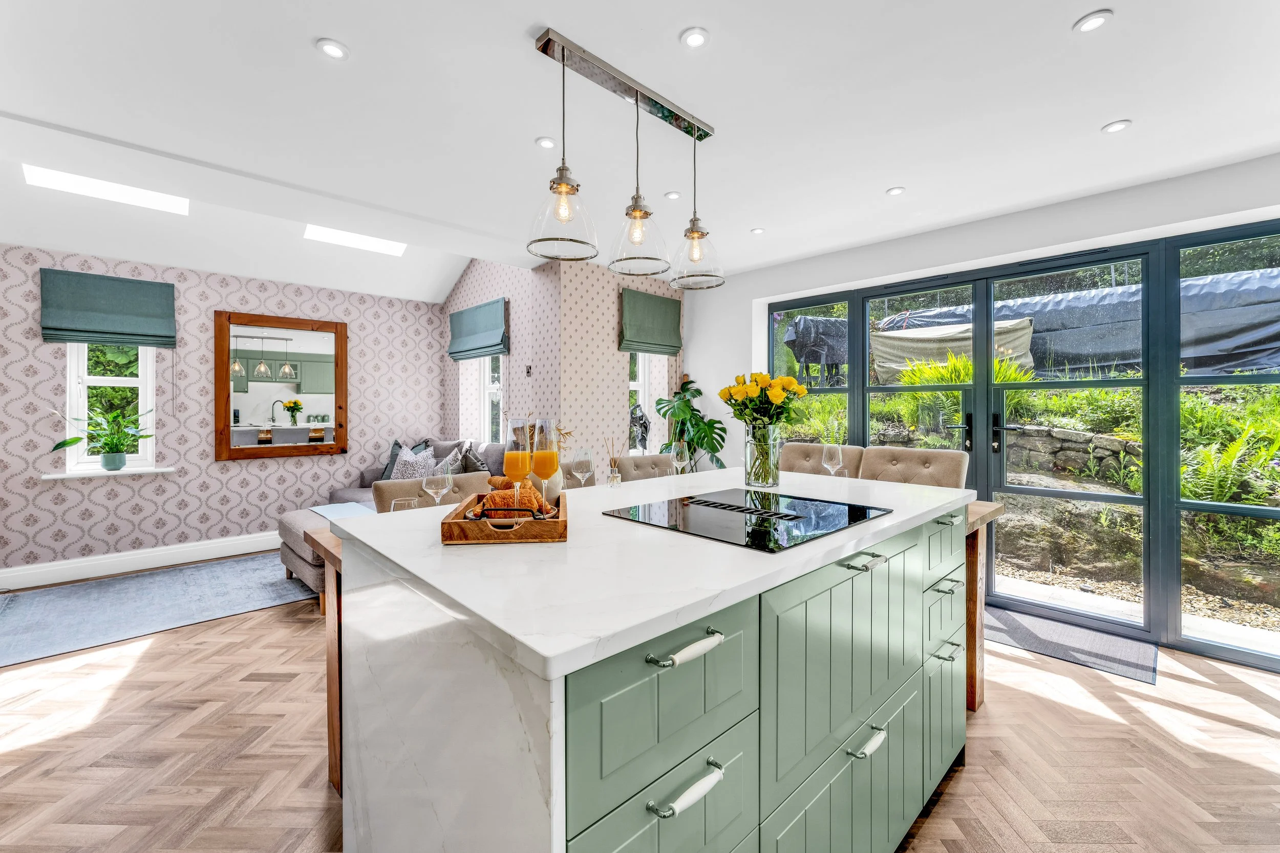 A bright, modern kitchen with large windows showing greenery outside. The kitchen has a white island with a green base, a black cooktop, and a vase with yellow flowers. In the background, there is a dining area with a beige bench, a table set with gl