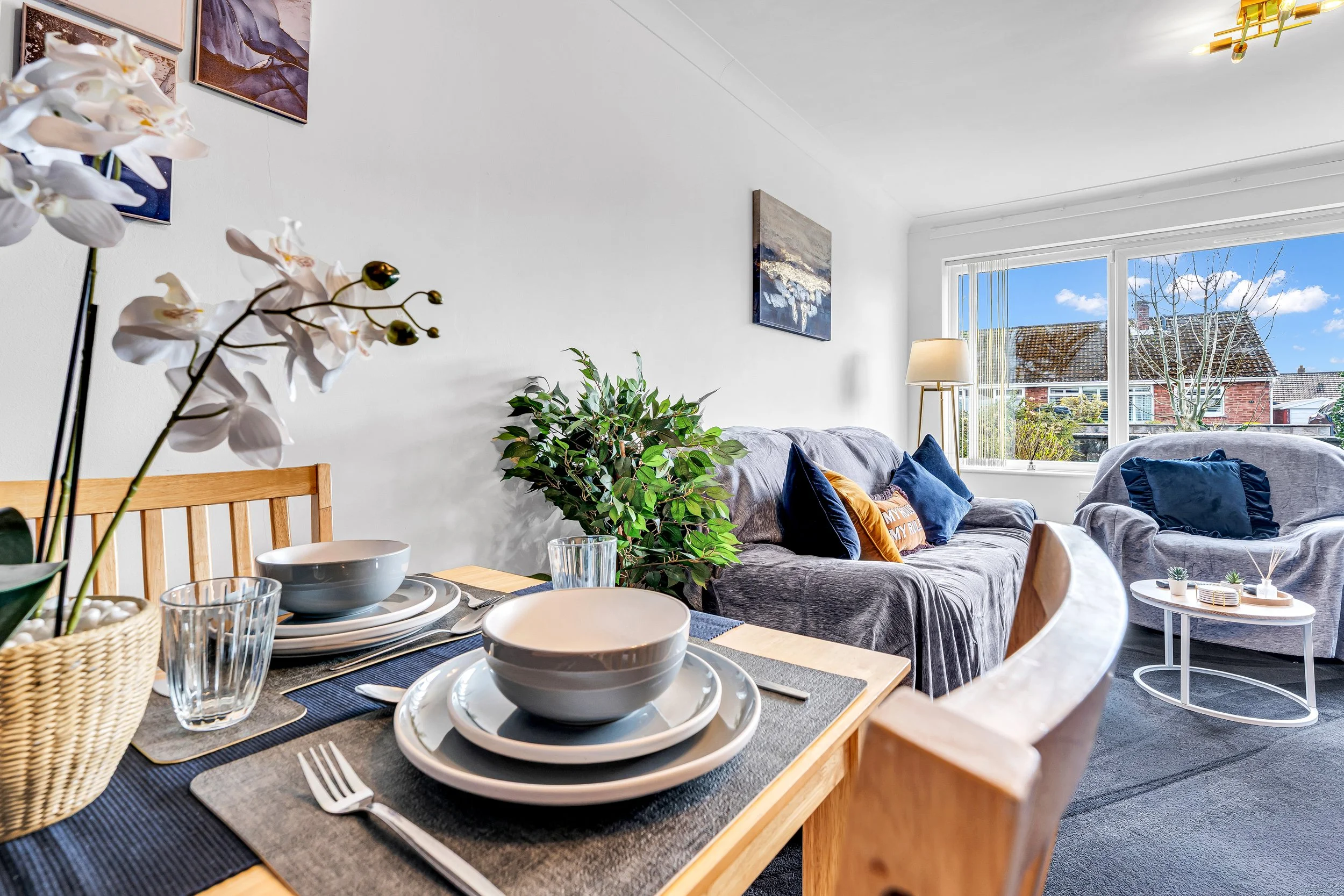 A cozy living room and dining area with a wooden table set with white and gray bowls, plates, glasses, and cutlery, decorated with a potted orchid plant. The living room has a gray sofa with colorful pillows, a white side table, a standing lamp, a la