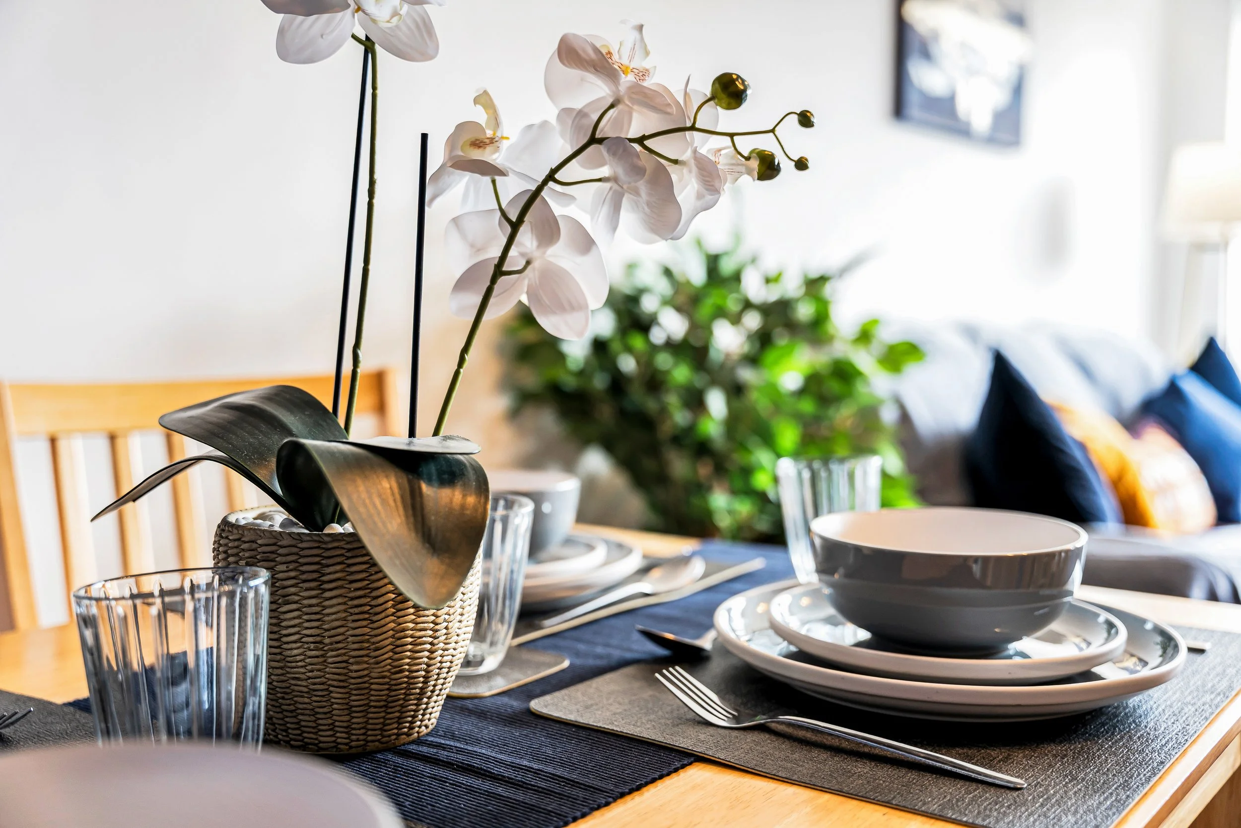 Dining table set with plates, bowls, utensils, and a potted orchid plant, with a sofa and houseplant in the background.