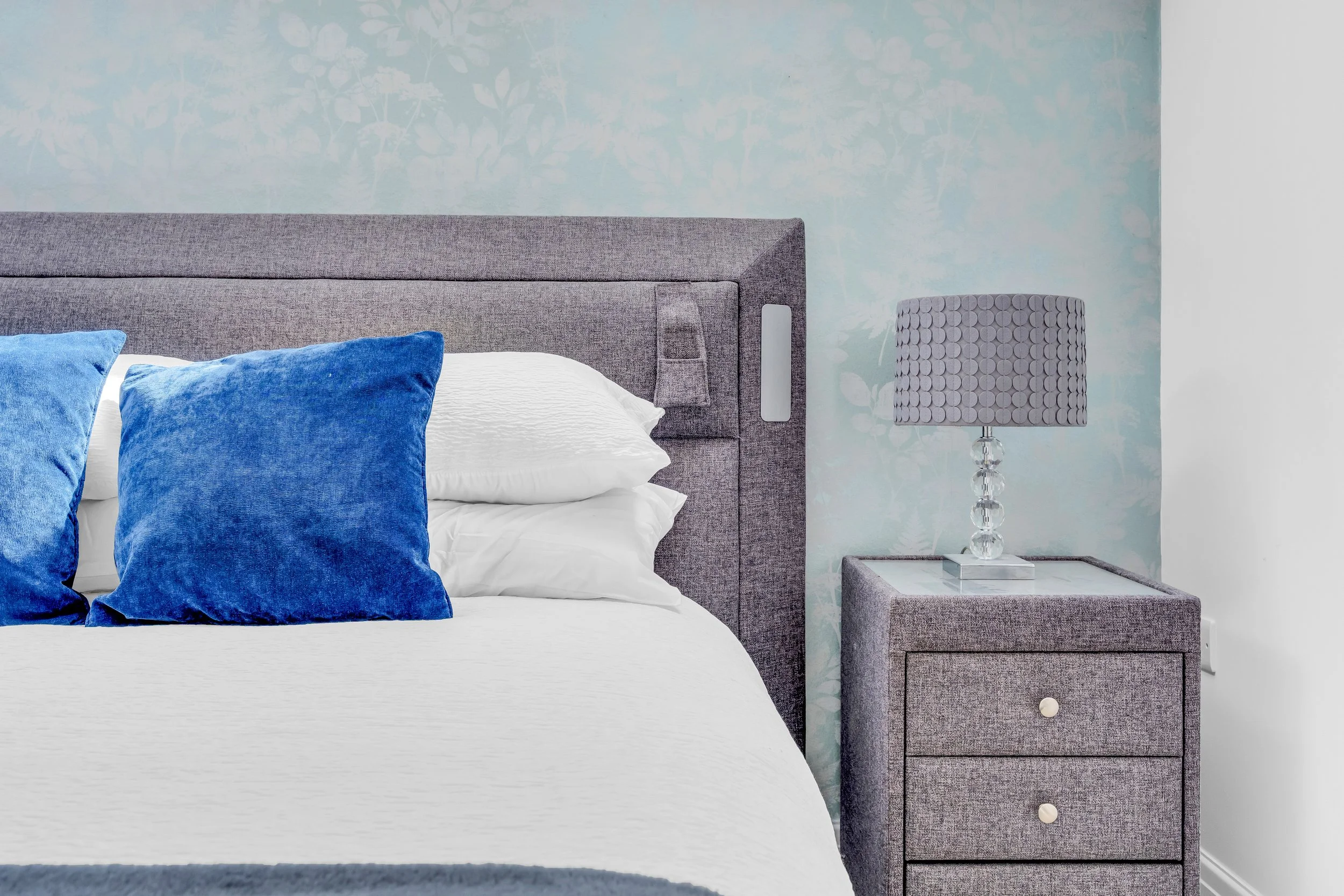 Close-up of a modern bedroom with a gray upholstered headboard, white bedding, and two blue velvet pillows. There is a matching gray nightstand with three drawers and a tall table lamp with a textured gray shade and a clear glass base. The wall behin