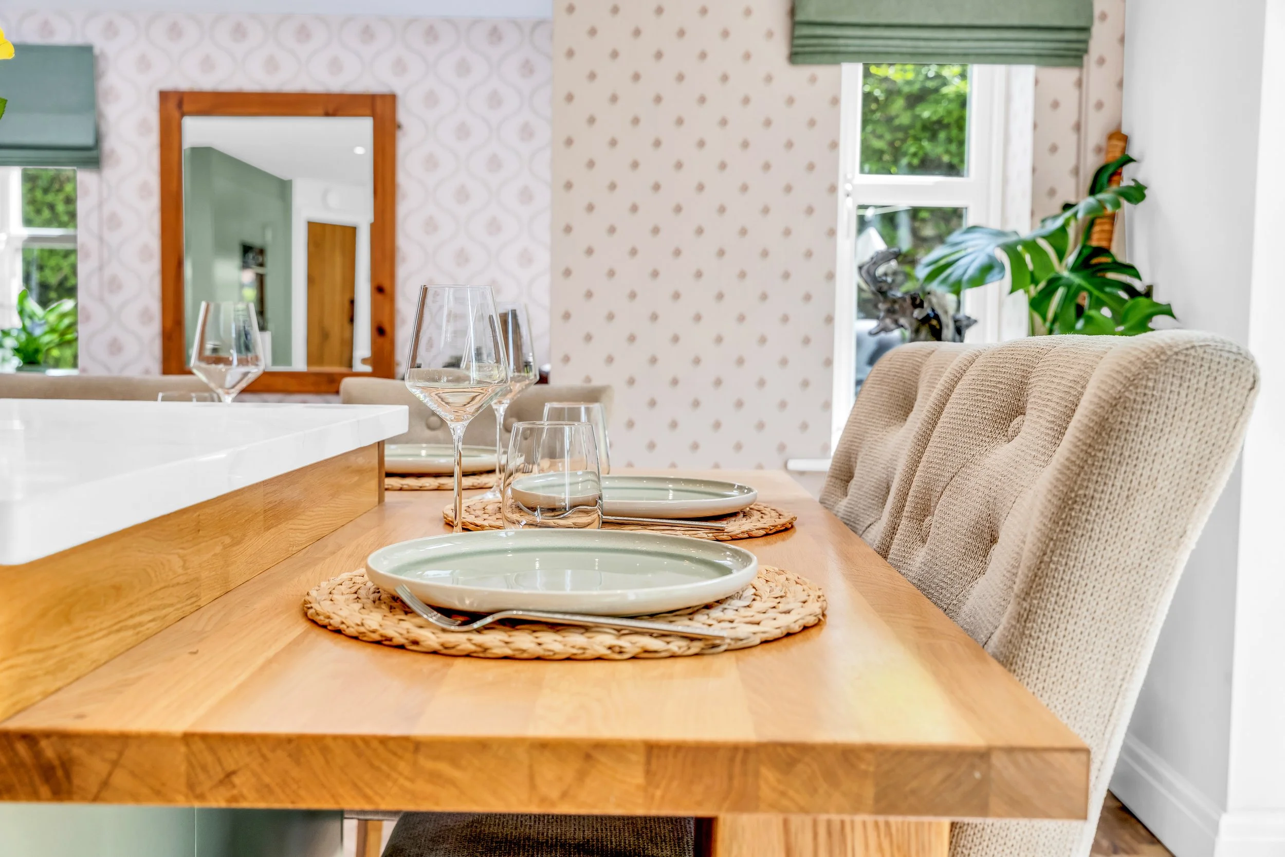 A dining room with a wooden table set with white plates, glassware, and woven placemats, surrounded by beige upholstered chairs, and a large window with a green valance showing greenery outside.