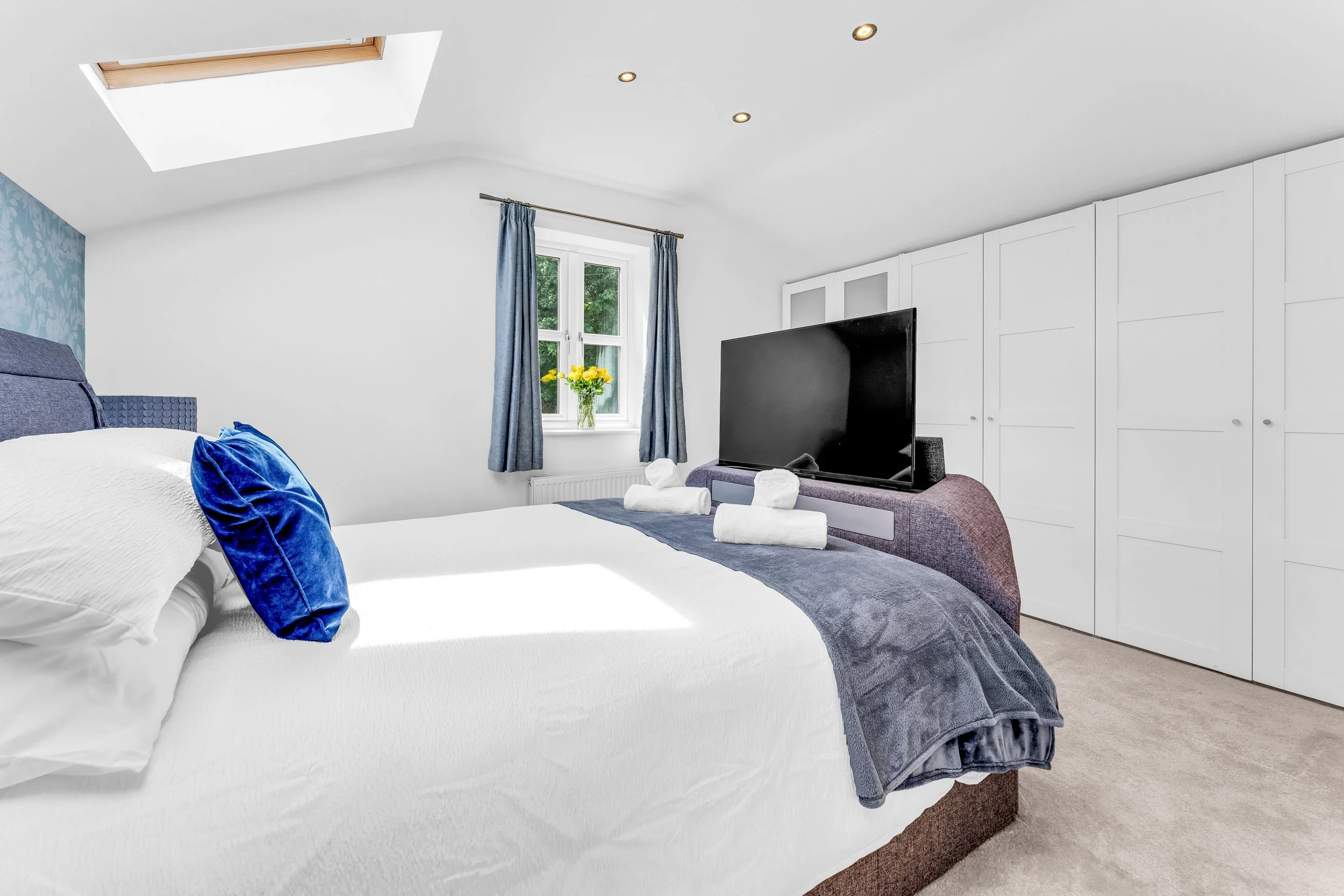 A bedroom with a white bed, blue pillows, a window with blue curtains, a TV on a purple bench, and yellow flowers on the windowsill.