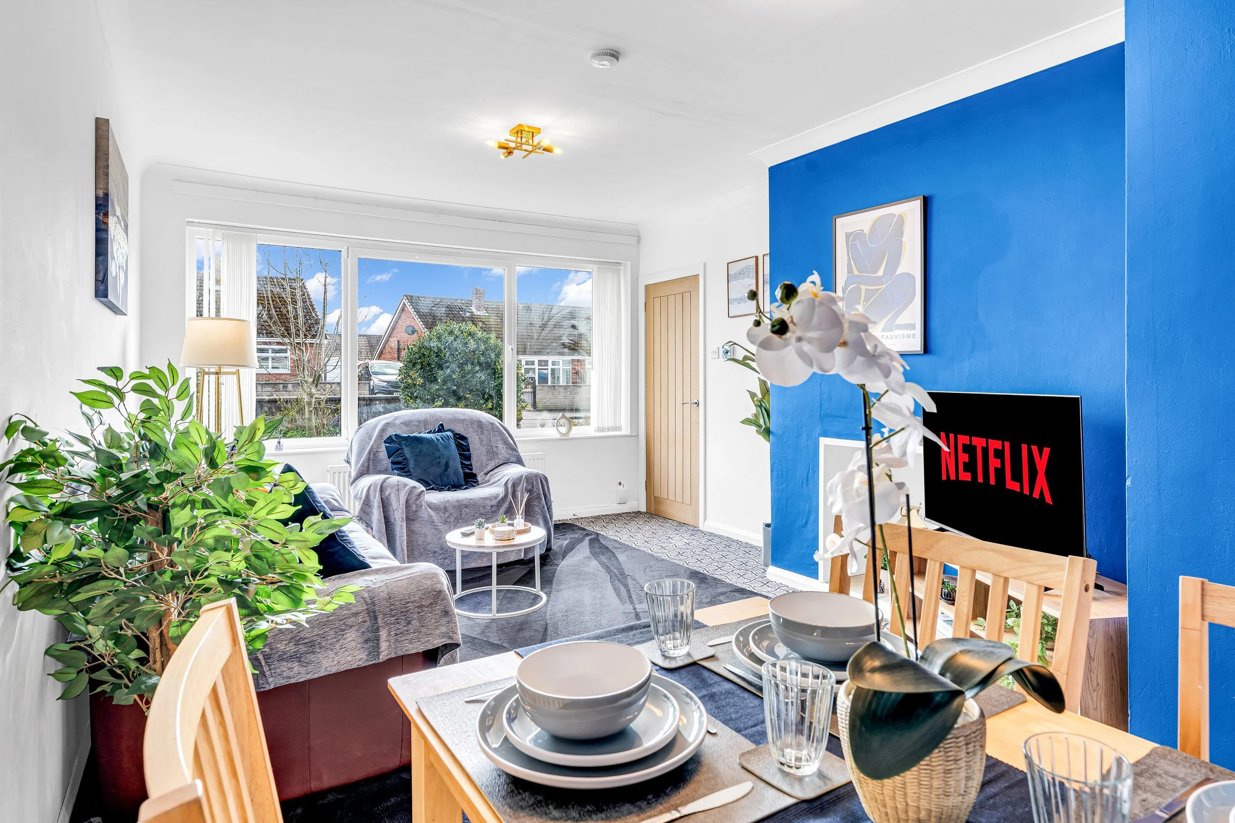 Bright living room with large window, blue accent wall, potted plant, dining table with place settings, and a TV displaying the Netflix logo.