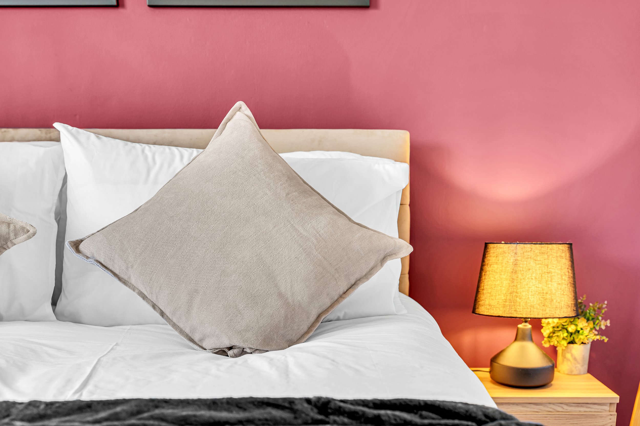 A bed with white sheets, beige pillows, and a wooden headboard against a pink wall. To the right, a bedside table with a yellow lamp and small potted yellow flowers.