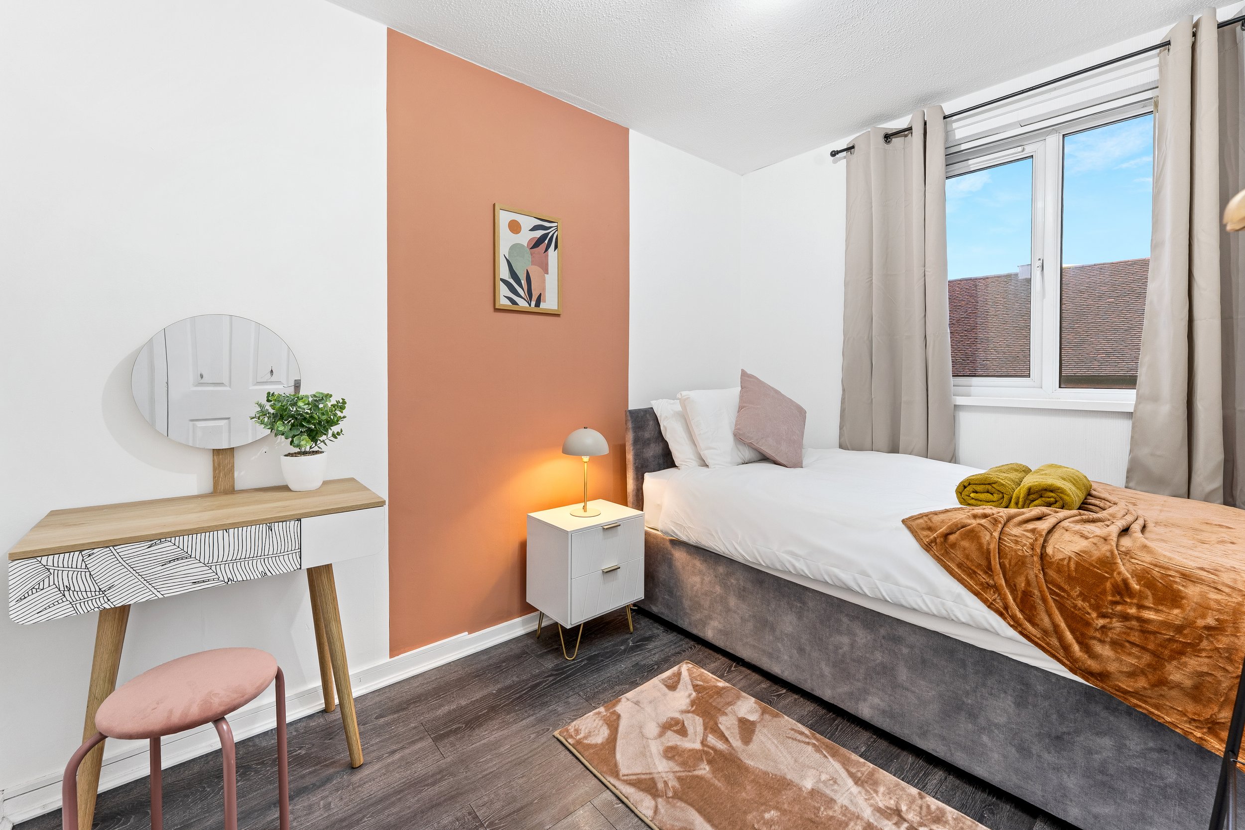 A cozy bedroom with a bed dressed in white and topped with yellow and pink blankets, a small white nightstand with a lamp, a window with beige curtains, a pink and beige rug, a vanity with a mirror and a small plant, and a framed abstract art piece on a peach-colored accent wall.