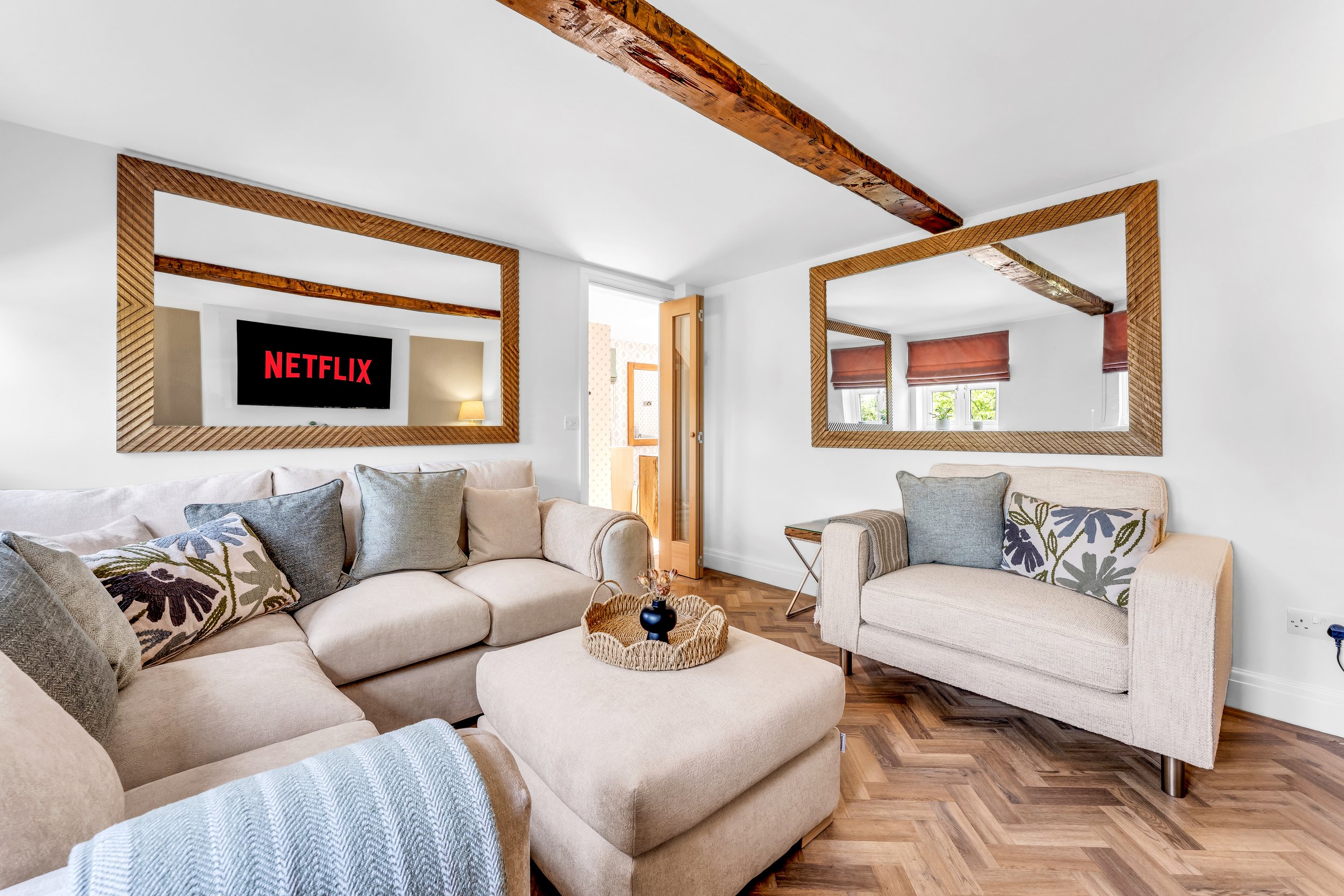 Living room with a large sectional sofa, patterned pillows, a coffee table with decor, and two large framed mirrors on the white walls. There are wooden beams on the ceiling and windows with red shades.