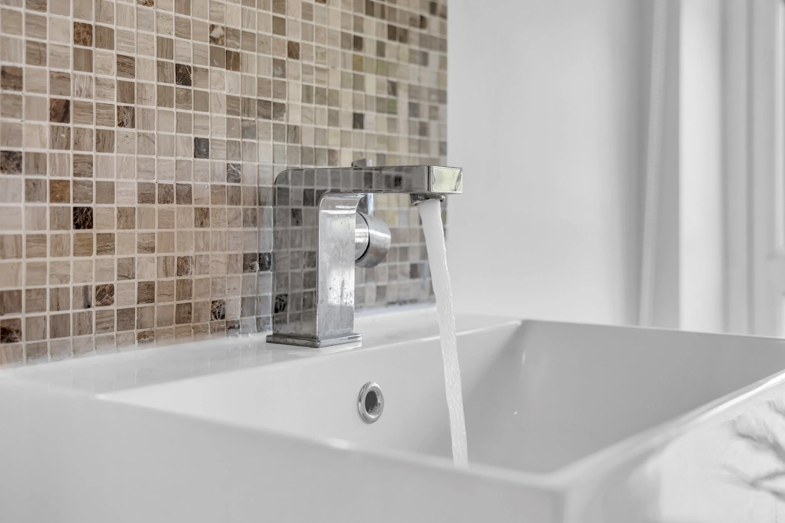 Close-up of a modern silver faucet with running water above a white sink, with a beige and brown mosaic tile backsplash.