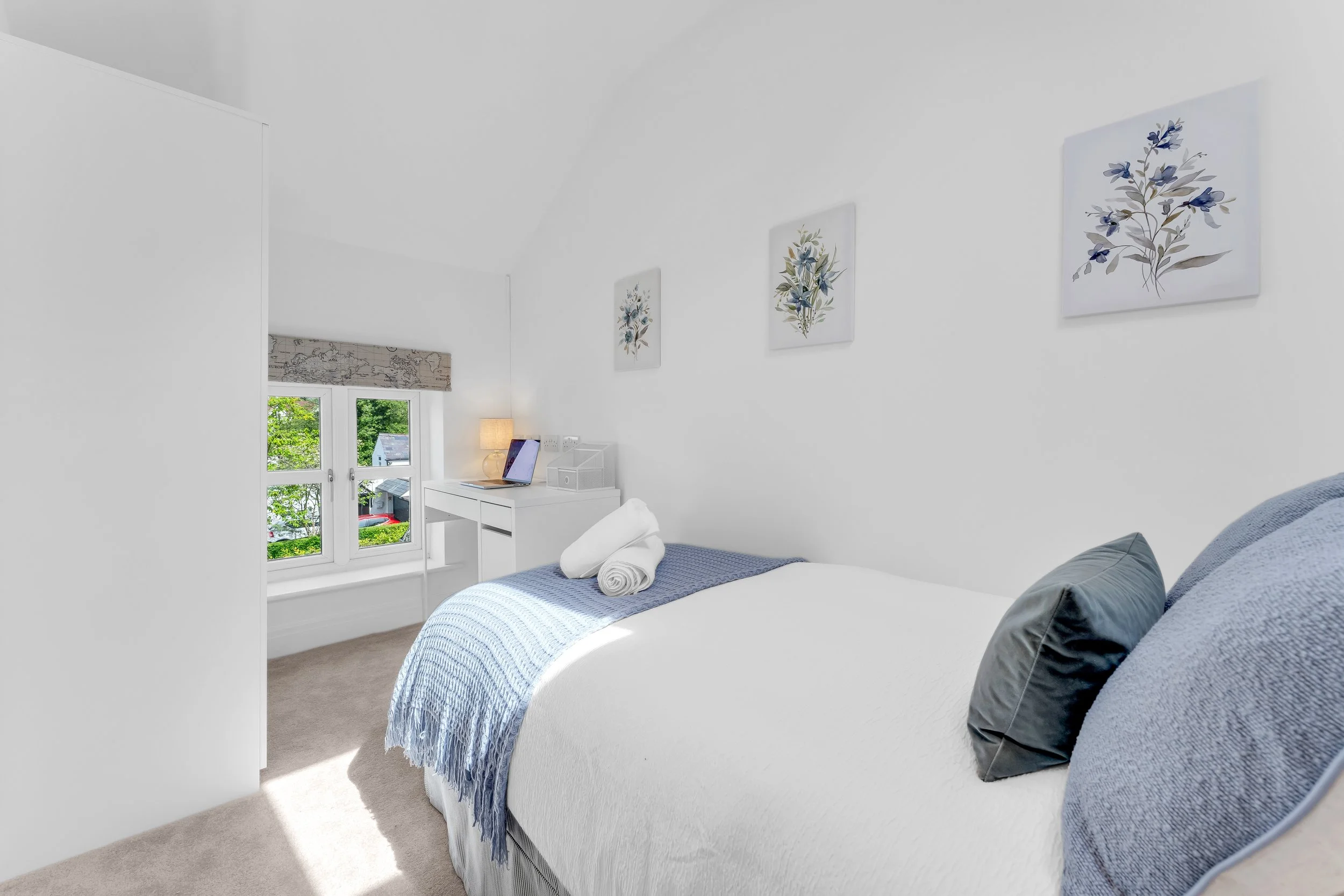 A bright, white bedroom with a bed covered in a light blue blanket and a gray pillow. There are three framed floral prints hanging on the wall and a window with a beige patterned roman shade. A white desk with a laptop, a lamp, and some storage conta