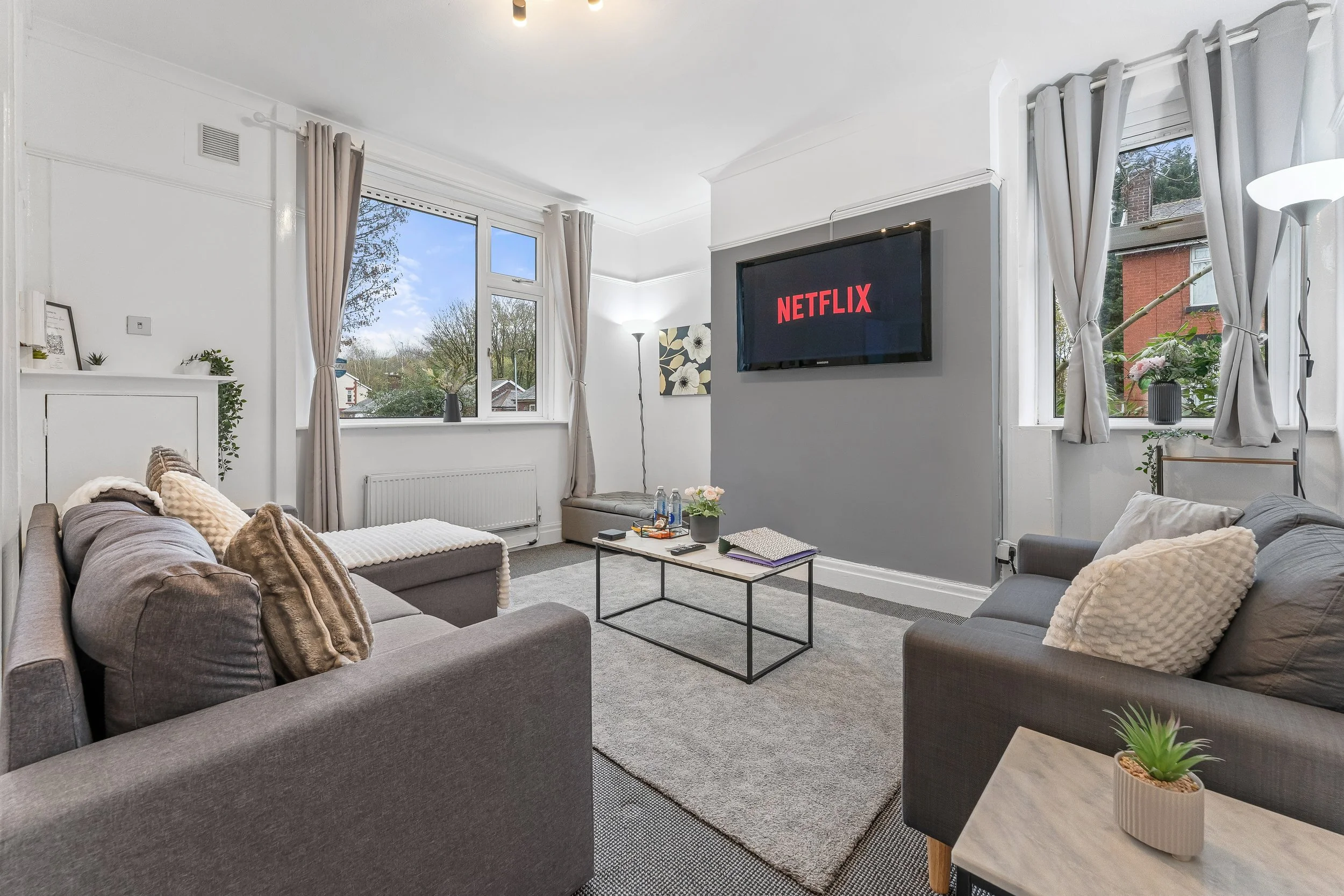 Living room with gray sofas, a TV displaying the Netflix logo, large windows with curtains, a coffee table with water bottles and notebooks, and decorative plants.