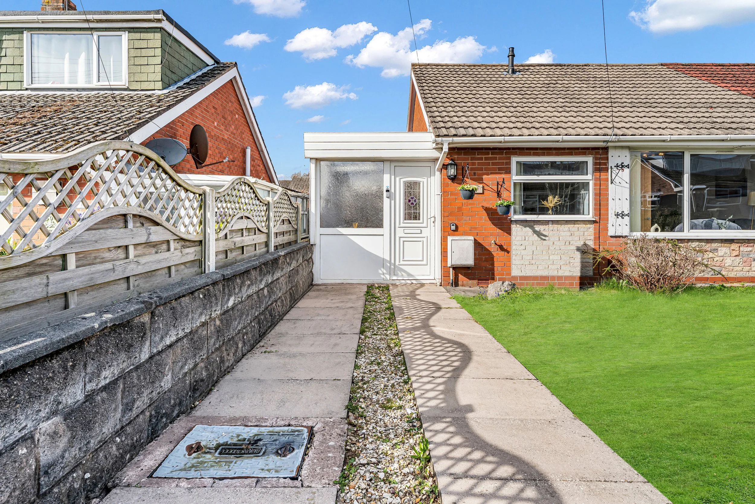 Front yard of a house with a concrete walkway leading to a white door with a small window, brick and white siding exterior, a lawn with bushes on the right, hanging planters and a lantern near the door, and a wooden fence on the left side.