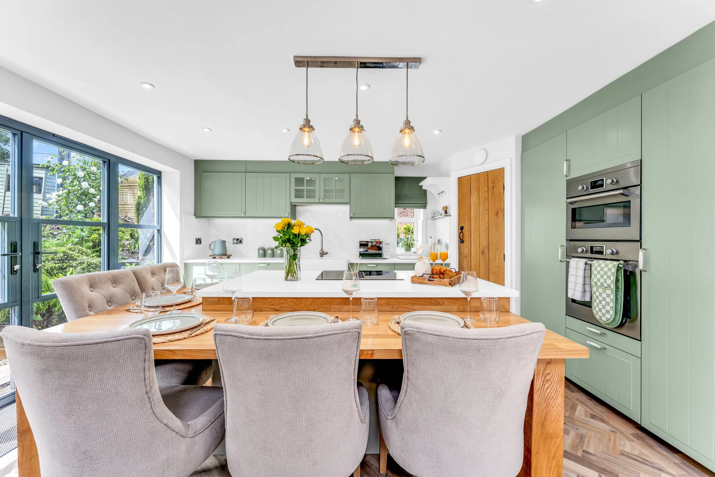 A bright, modern kitchen with green cabinets, a white island, and a wooden dining table with gray upholstered chairs, decorated with a vase of yellow flowers and tableware.