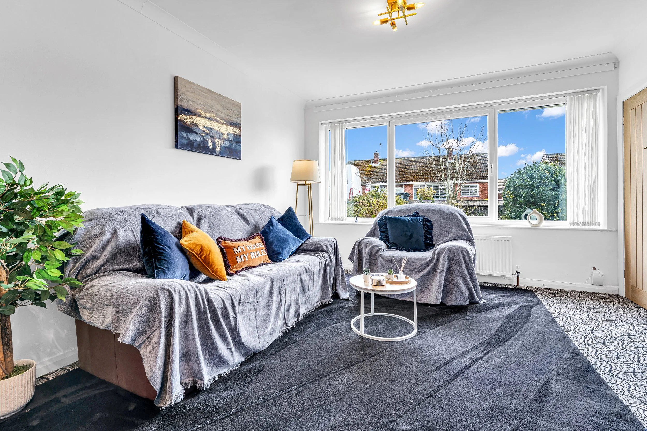 Living room with gray sofa and armchair, colorful pillows, a white round coffee table, large window with a view of trees and houses, potted plant, floor lamp, artwork on white wall, contemporary decor.