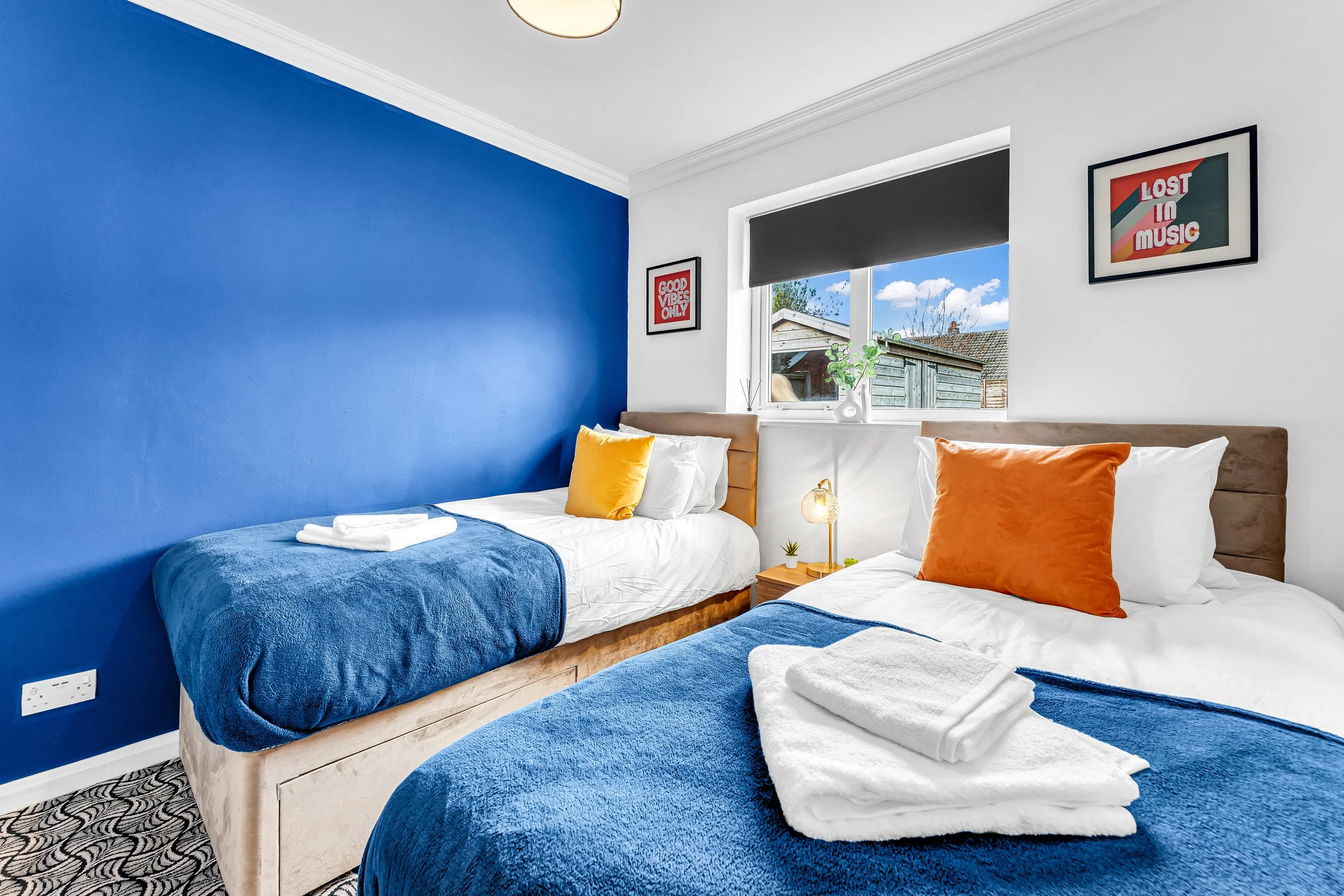 A bedroom with two beds, one with a blue headboard and bedding, the other with a beige headboard and matching bedding, a window with a view outside, wall decorations, and neatly folded towels on each bed.