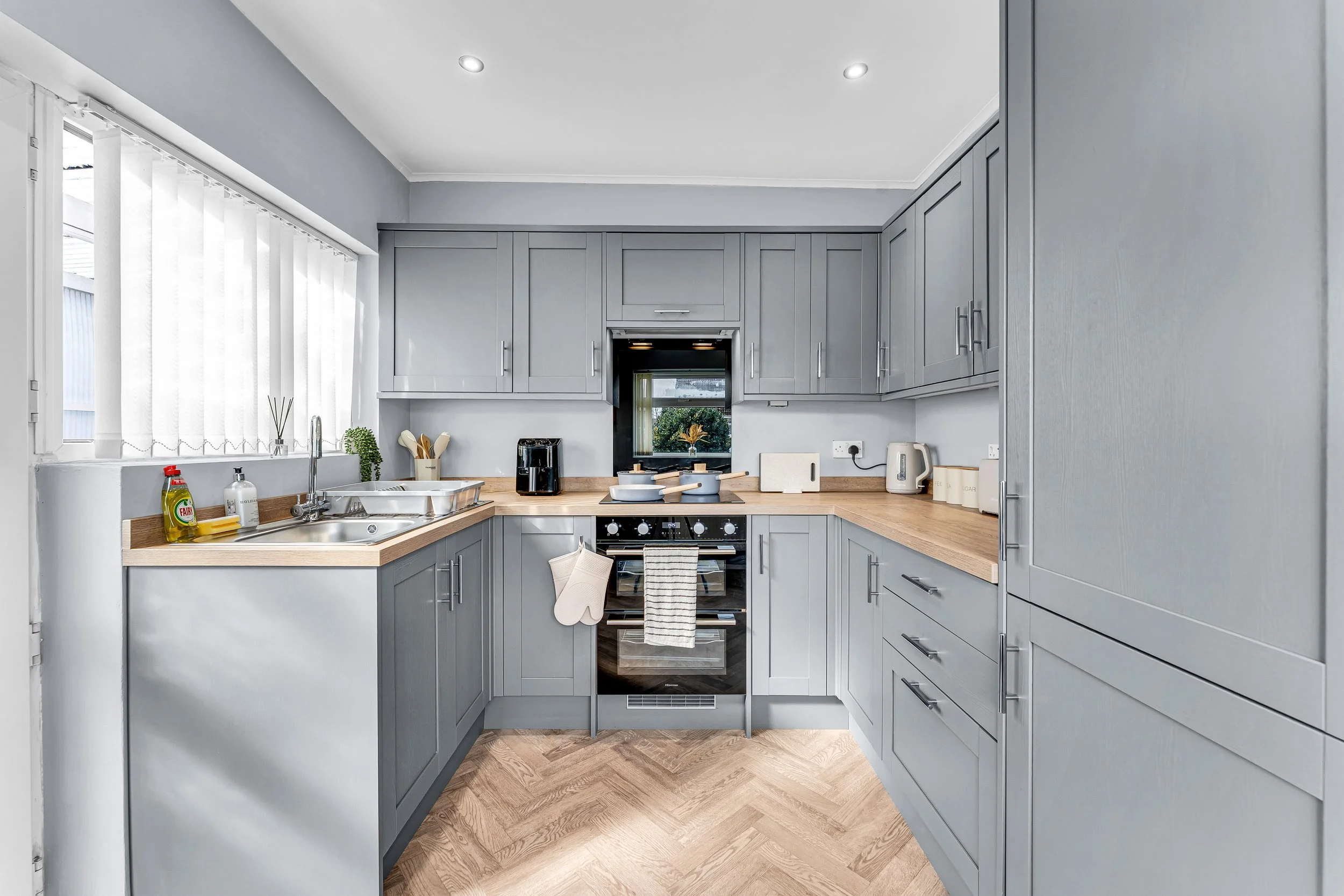 Modern kitchen with grey cabinets, wooden countertops, and a window with vertical blinds, featuring kitchen appliances and utensils.