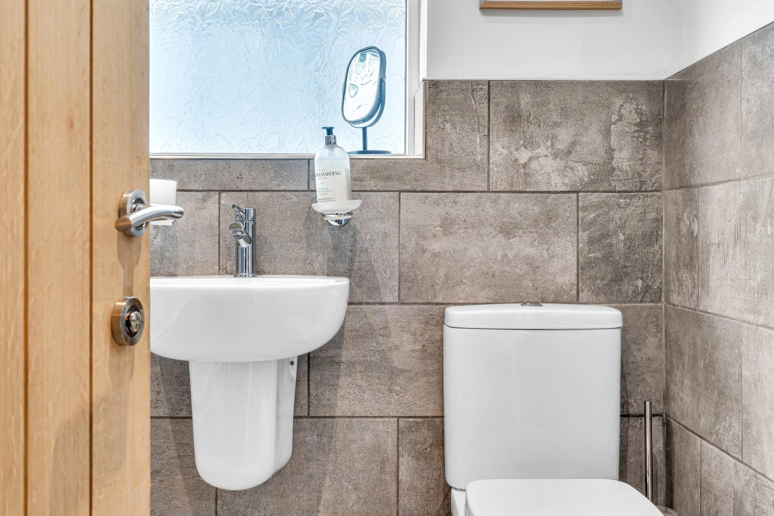 A small bathroom with tiled gray walls, a white toilet, a white wall-mounted sink with a chrome faucet, a small mirror, and a window with frosted glass. A hand soap dispenser is mounted on the wall above the sink.