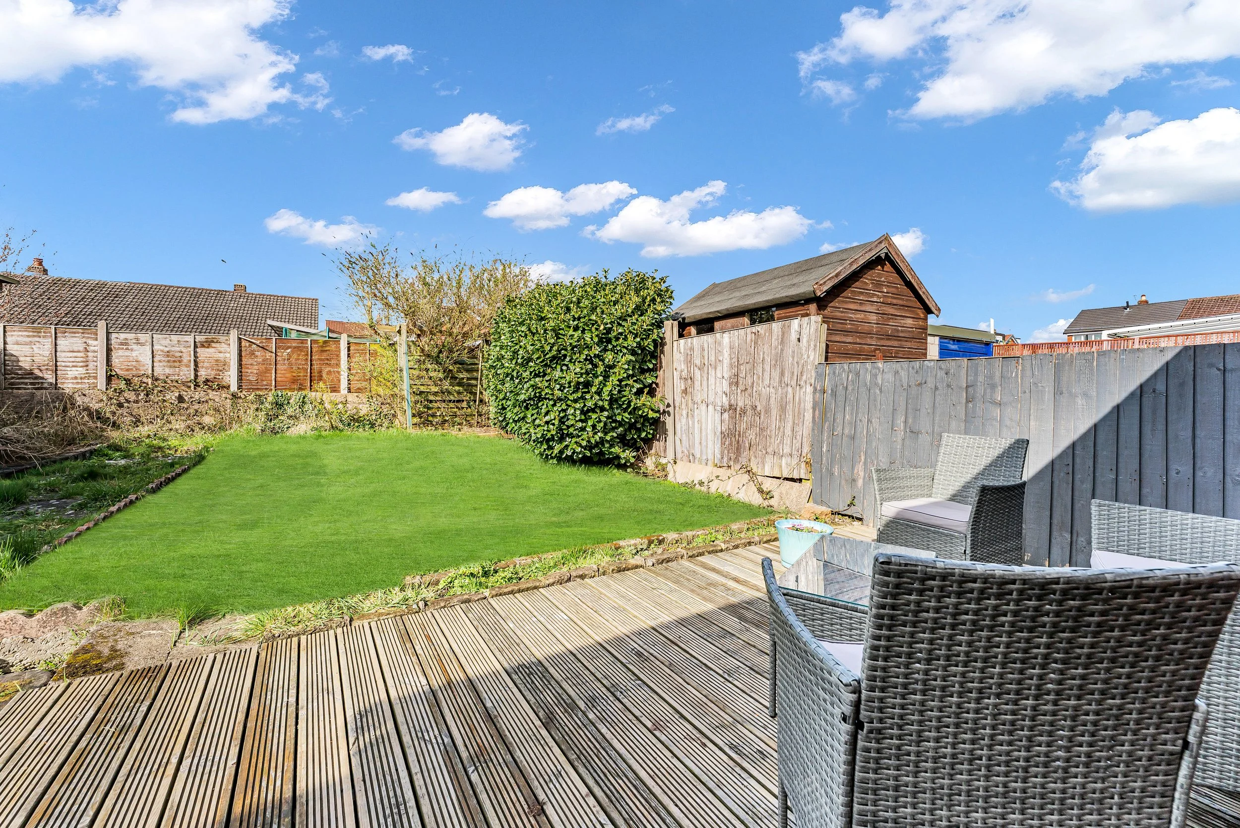 Backyard with a wooden deck, outdoor chairs, a grassy lawn, a bush, and a wooden shed under a blue sky with clouds.