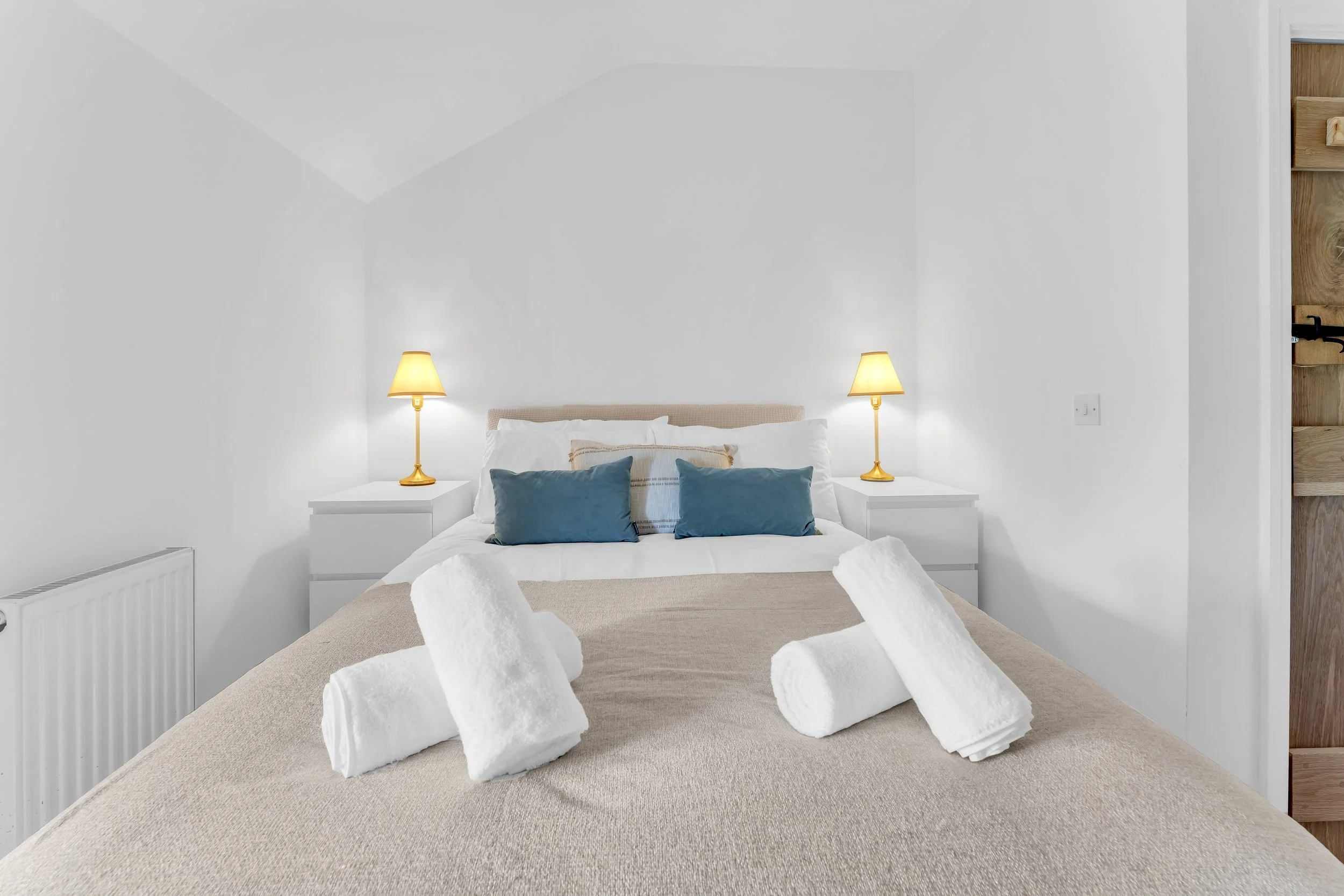 A neatly made bed with beige bedding and four pillows, two white pillows and two blue pillows, with towel swans arranged on the bed in front of the pillows. There are two yellow bedside lamps on white nightstands on either side of the bed, with a whi