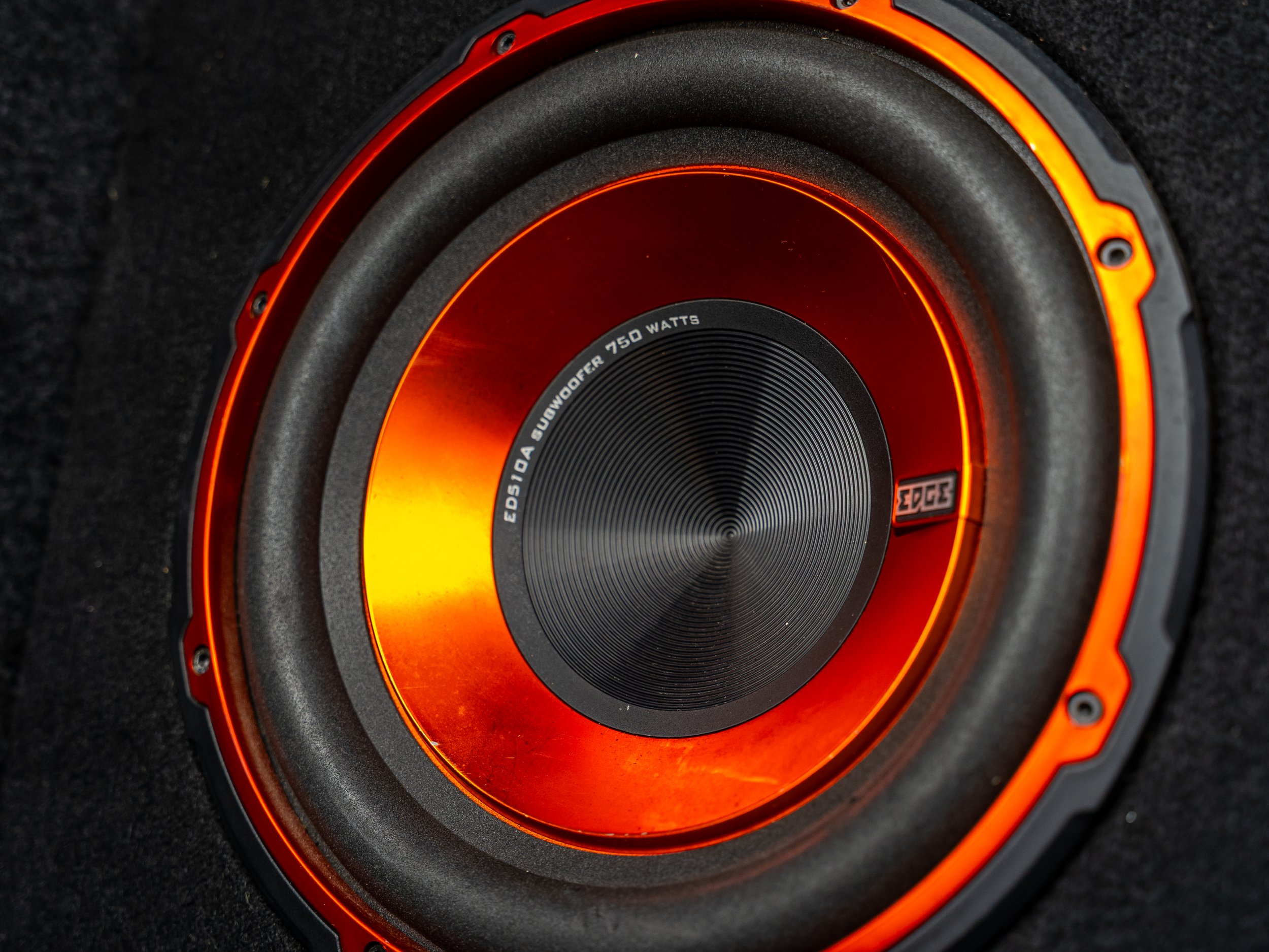 A close-up of an orange and black subwoofer speaker with the brand and specifications visible.