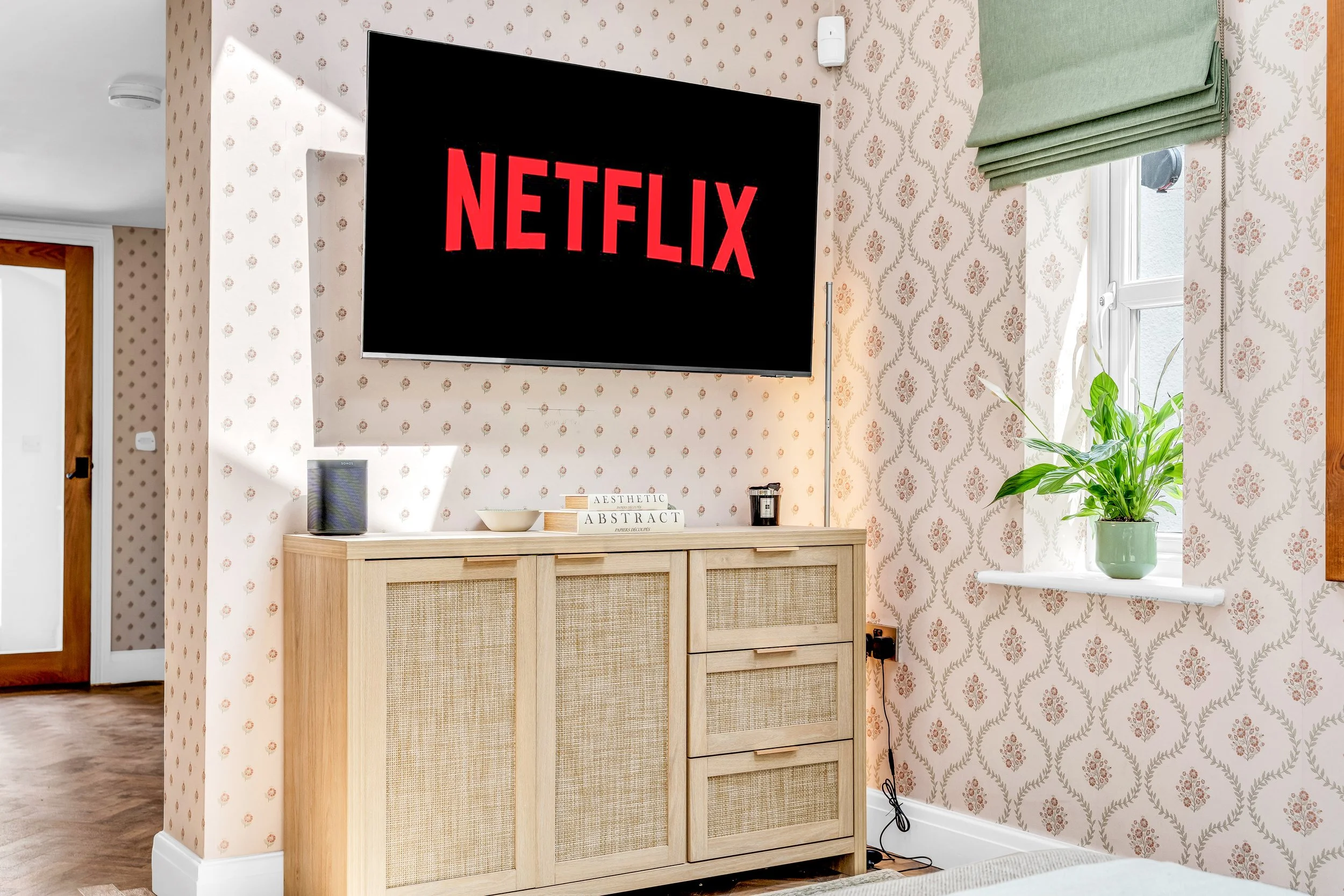 A living room with patterned wallpaper, a mounted flat-screen TV displaying the Netflix logo, a light wood cabinet with rattan panel doors, a potted plant on a white shelf near a window with green blinds, and decorative books and a speaker on the cab