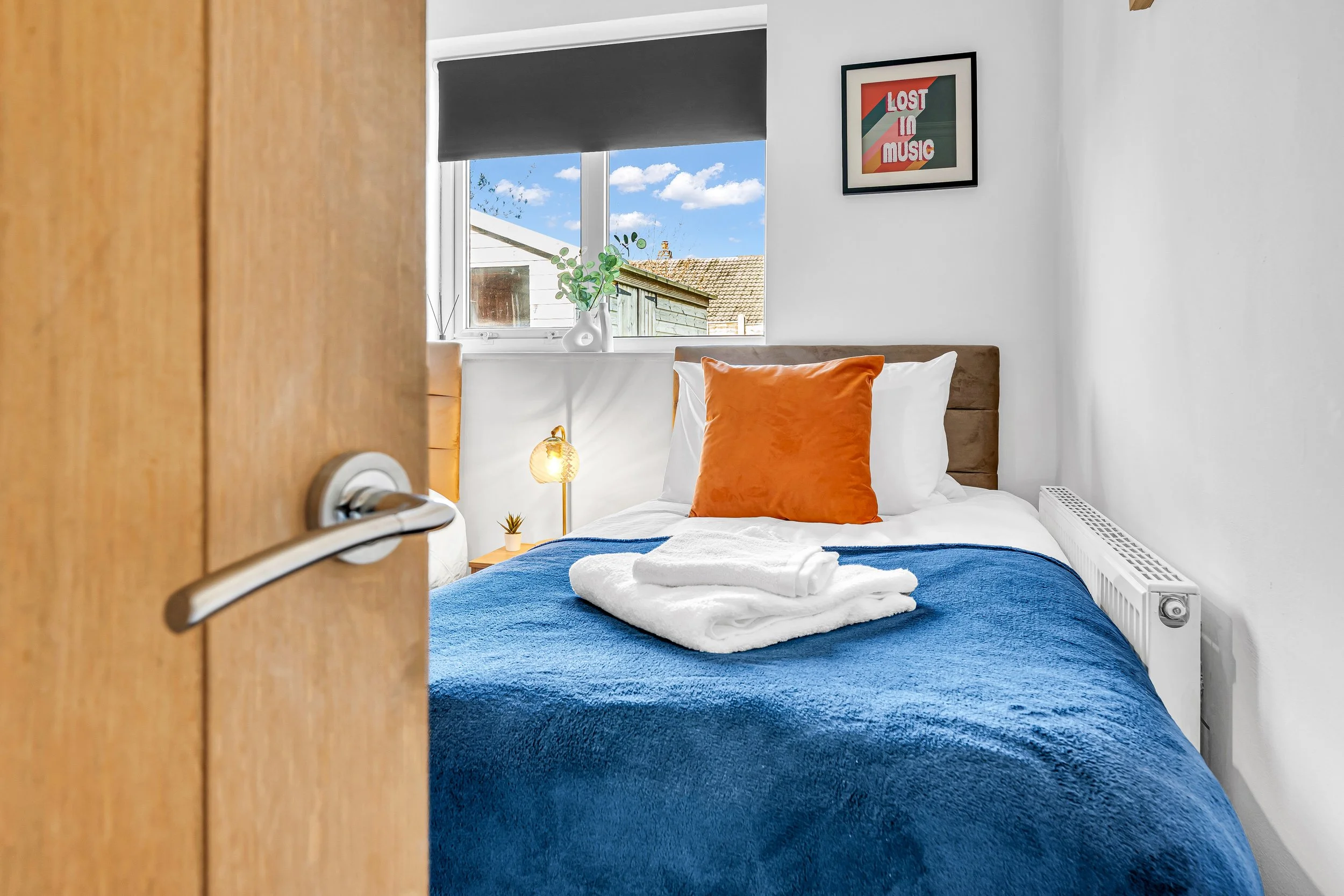 A cozy bedroom with a peek through a partially open wooden door. The bed has white linens, a blue blanket, and an orange pillow. Towels are neatly folded on top of the bed. A bedside table with a small potted plant and a lamp is visible. There's a wi